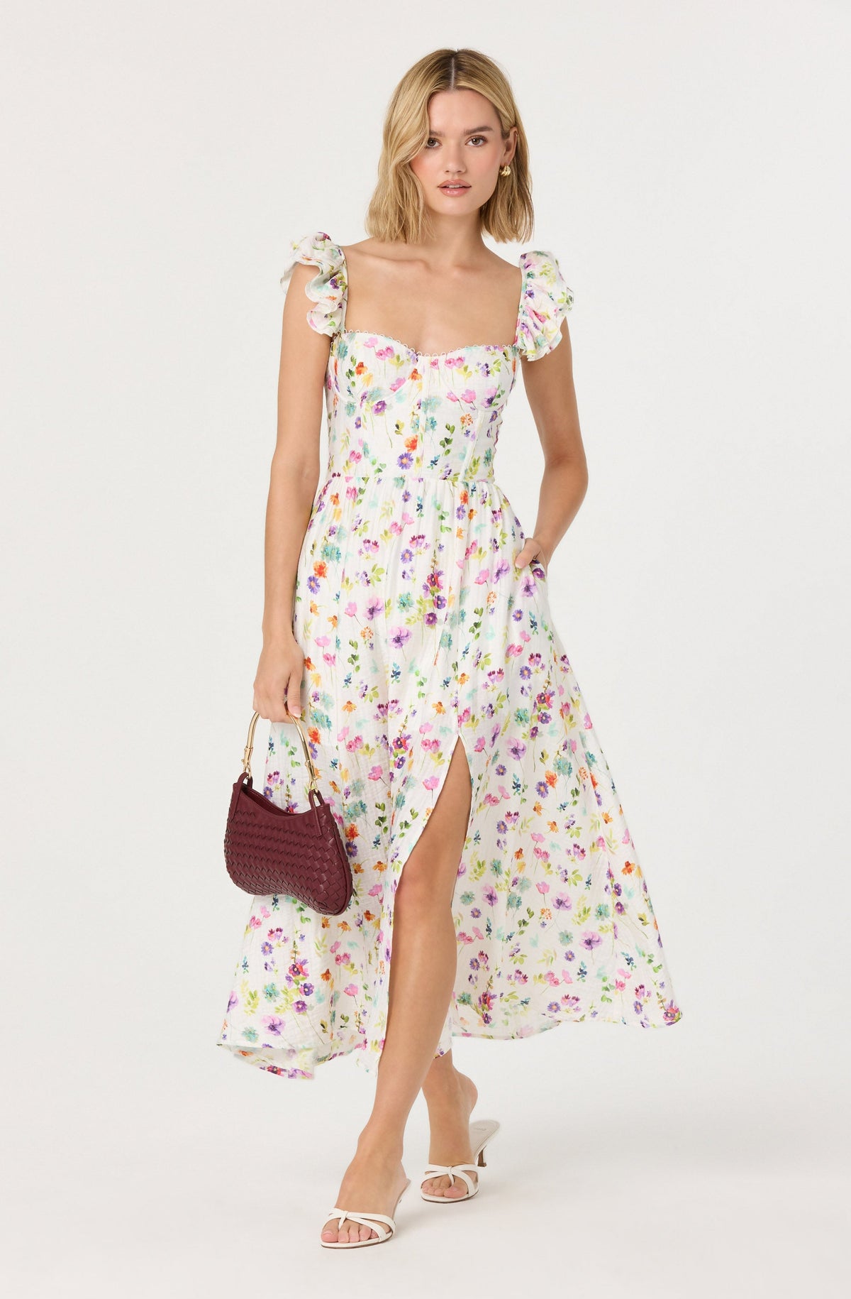 A woman is wearing a long, sleeveless, floral dress with ruffled shoulder straps. The dress has a sweetheart neckline, a fitted waist, and a front slit. She is holding a dark red woven handbag with a gold handle and is wearing white, open-toe high-heeled shoes.