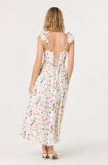 A woman is wearing a long, flowing dress with a colorful floral print on a white background. The dress features short ruffled sleeves and a lace-up back with crisscrossed laces. She is also wearing a pair of white high-heeled shoes.