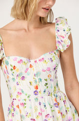 A woman is wearing a sleeveless dress with a floral pattern on a white background. The dress has ruffled, short sleeves and a fitted bodice with visible vertical seams. The floral print features various colors, including pink, purple, orange, green, and blue, with small, delicate flowers. The dress has a peach-colored, gathered waistline. The material appears light and airy, perfect for warm weather.