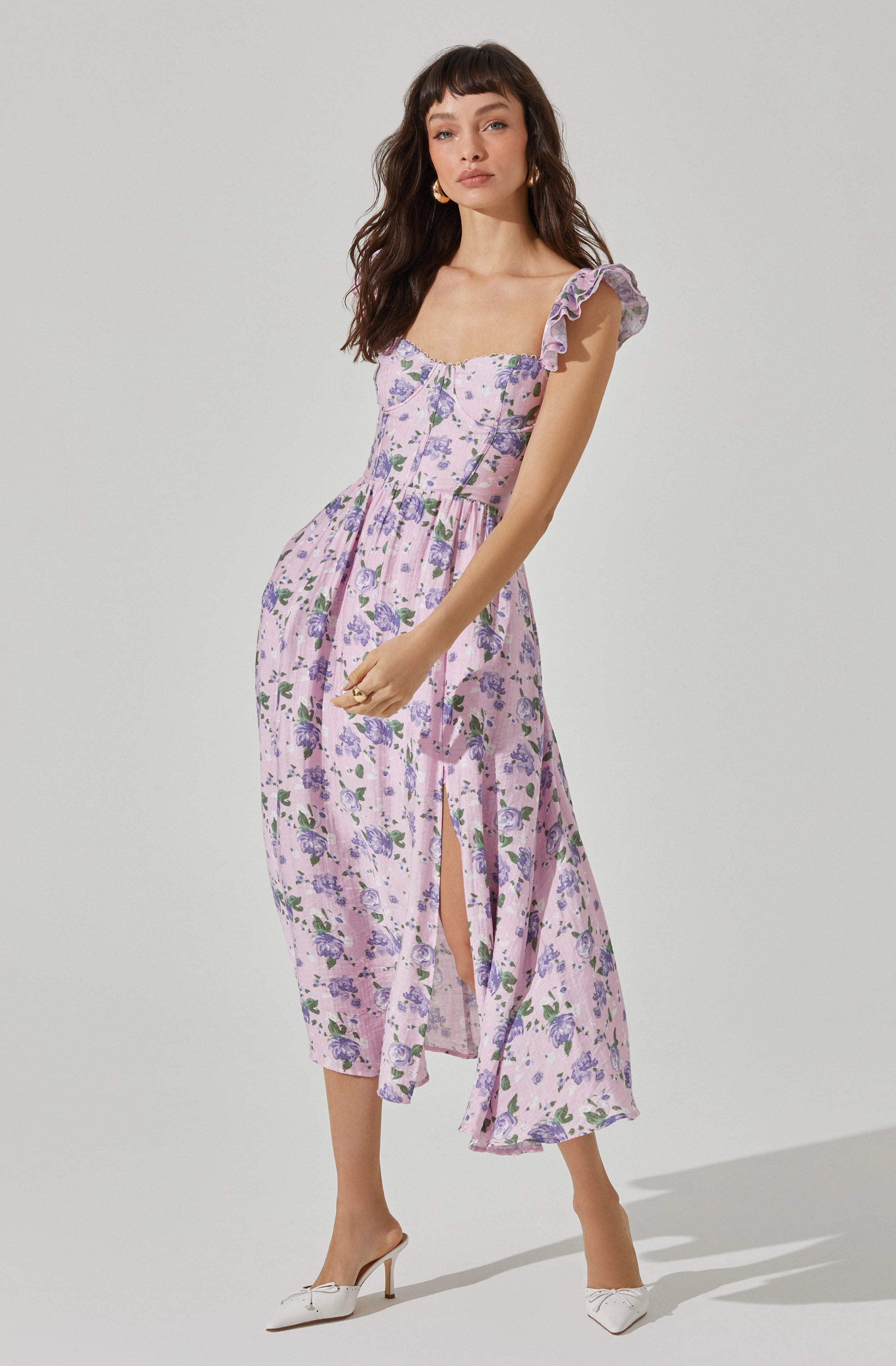 A woman is wearing a light pink, floral dress with purple and green flowers. The dress has ruffled sleeves, a fitted bodice, and a slit on the side that reaches above her knee. She is also wearing white high-heeled shoes.