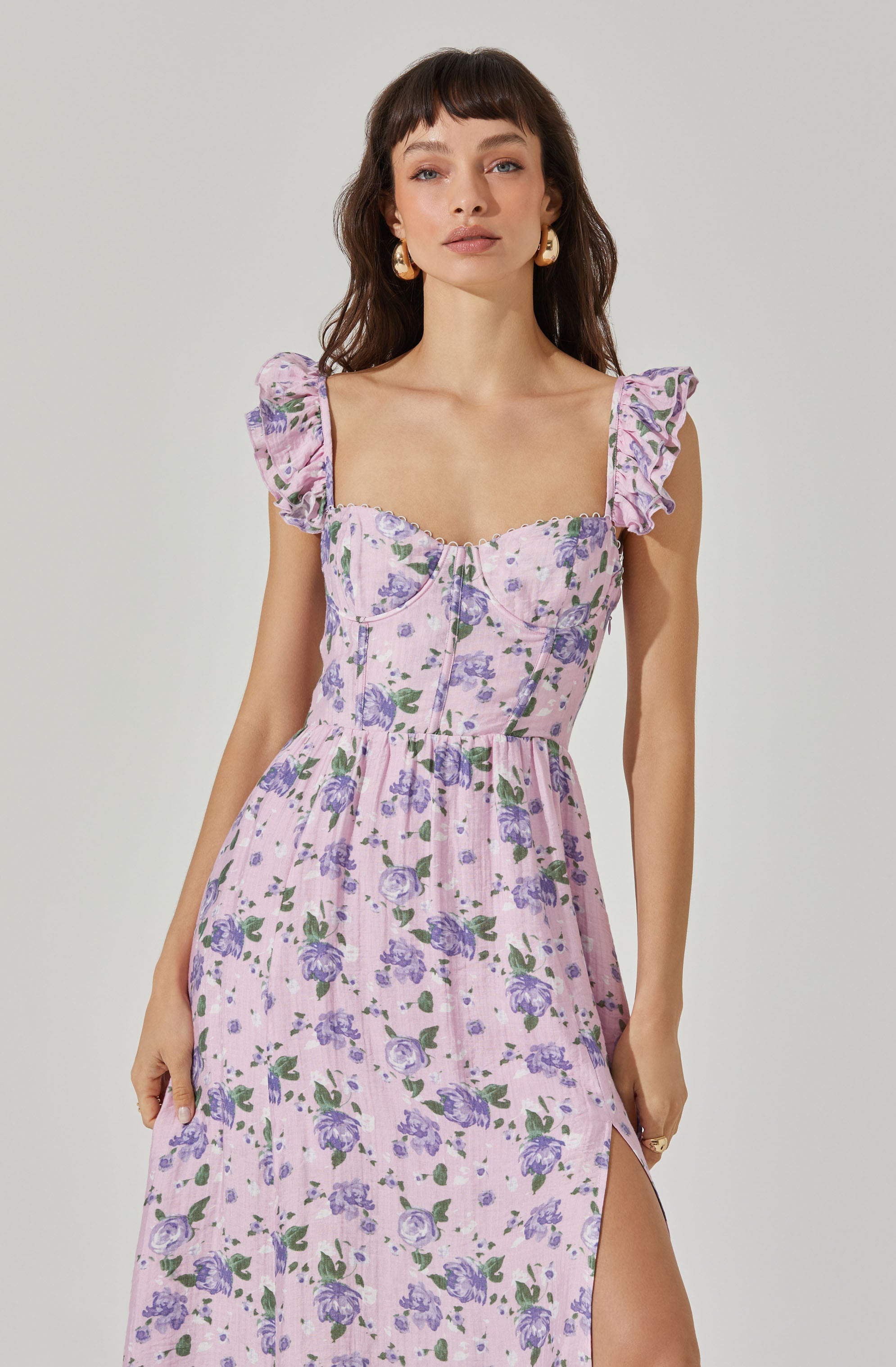 A woman is wearing a light pink dress with a purple floral pattern. The dress features puffed, ruffled short sleeves with a floral design, a fitted bodice with sweetheart neckline and visible seaming details, and a flowing skirt with a thigh-high slit on the left side.
