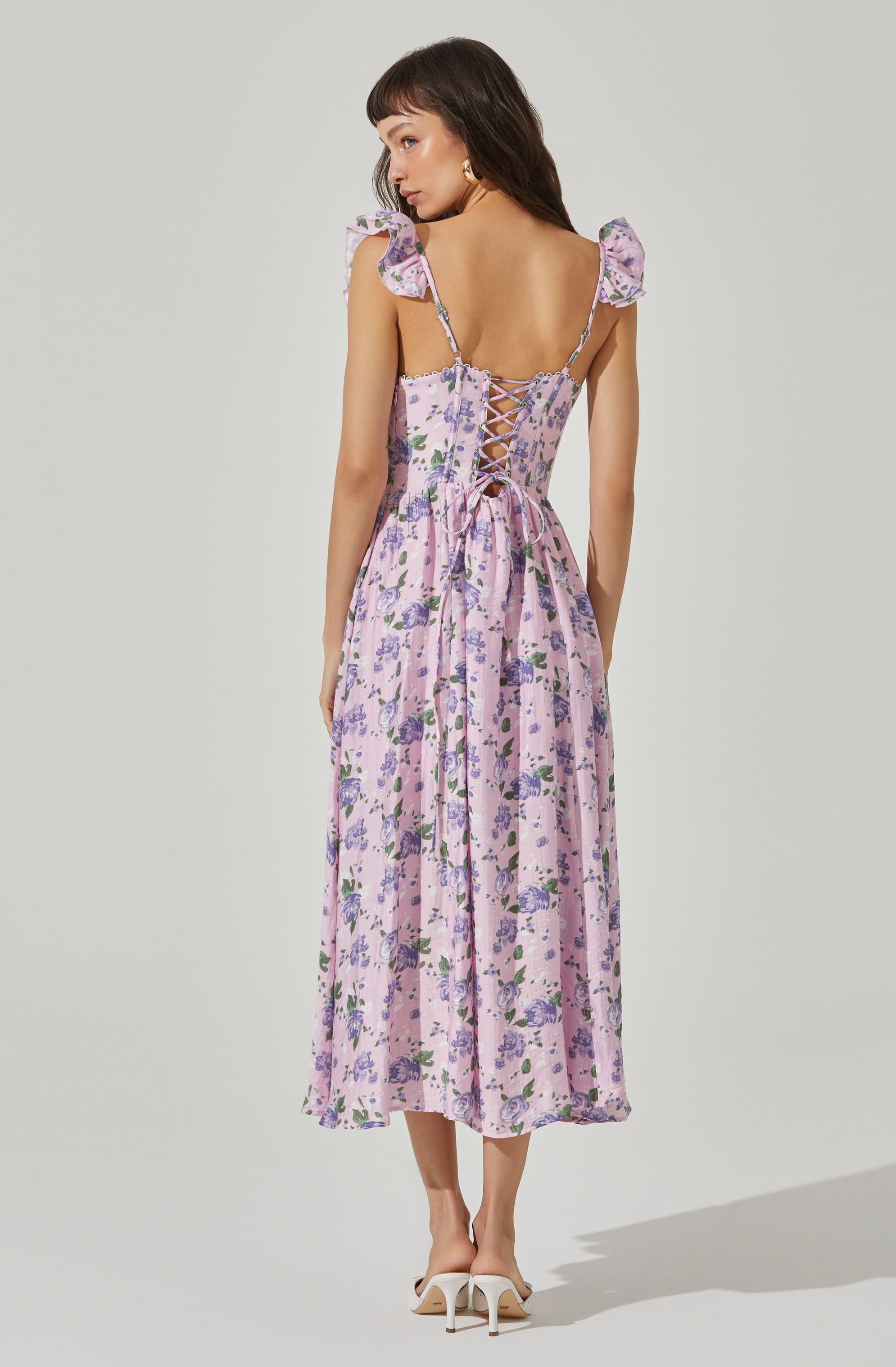 A woman wearing a long, flowy, pink dress with a purple floral pattern. The dress has ruffled shoulder straps and a lace-up back detail with thin ties. She is also wearing white high-heeled shoes.