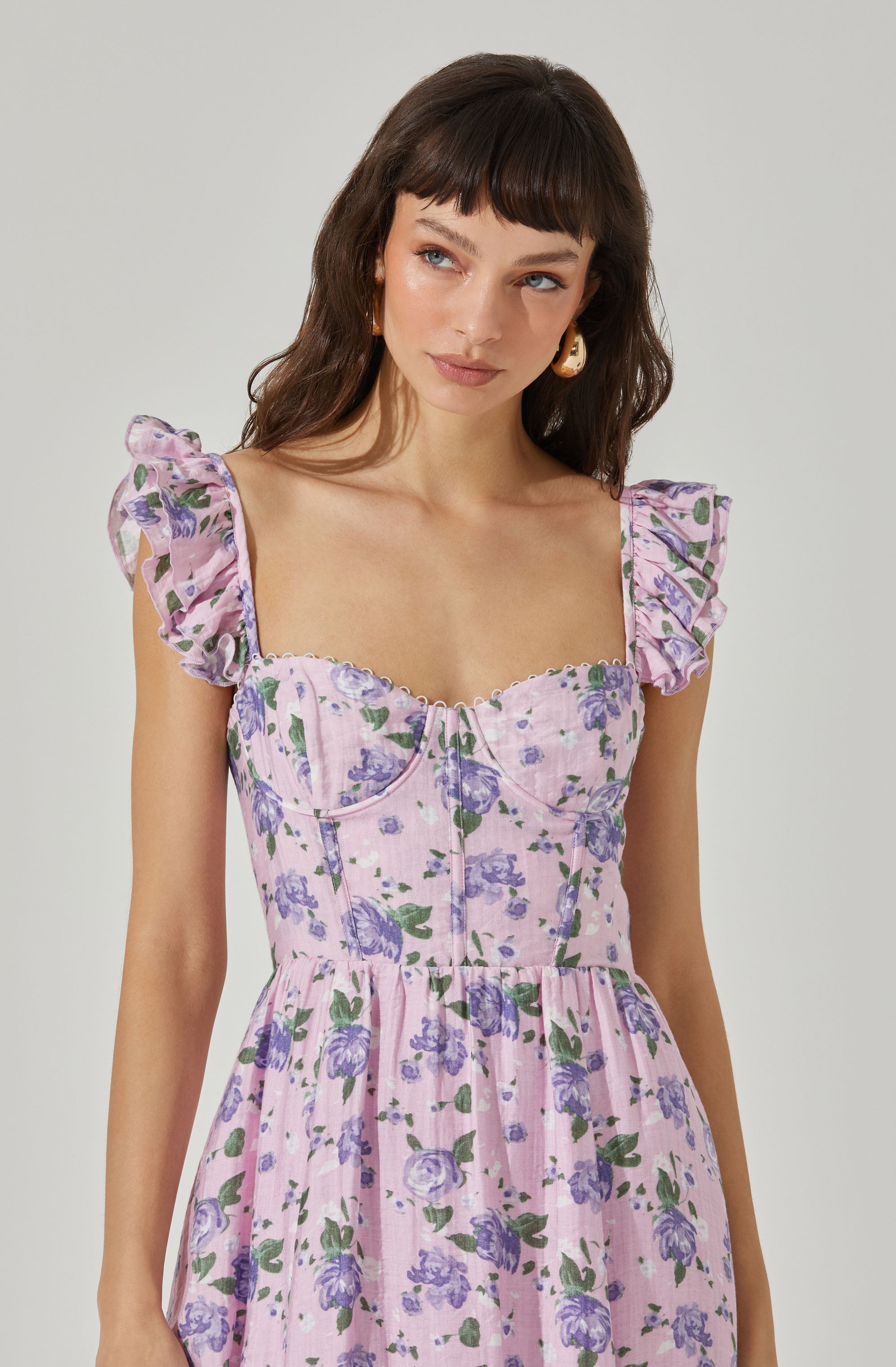 A woman wears a pink dress with a purple floral pattern, featuring ruffled cap sleeves and a fitted bodice with visible paneling and seam details.