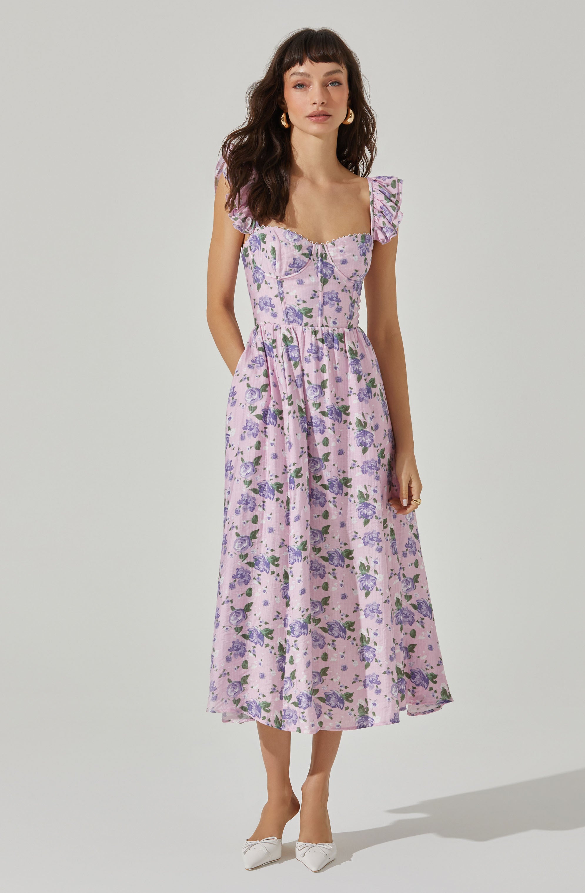A woman is wearing a pink floral dress with purple and green flowers. The dress features puffed cap sleeves with ruffles, a fitted bodice with a sweetheart neckline, and a full, flowing skirt that falls to mid-calf. She is accessorizing with gold hoop earrings and white pointed-toe shoes with a small bow detail.
