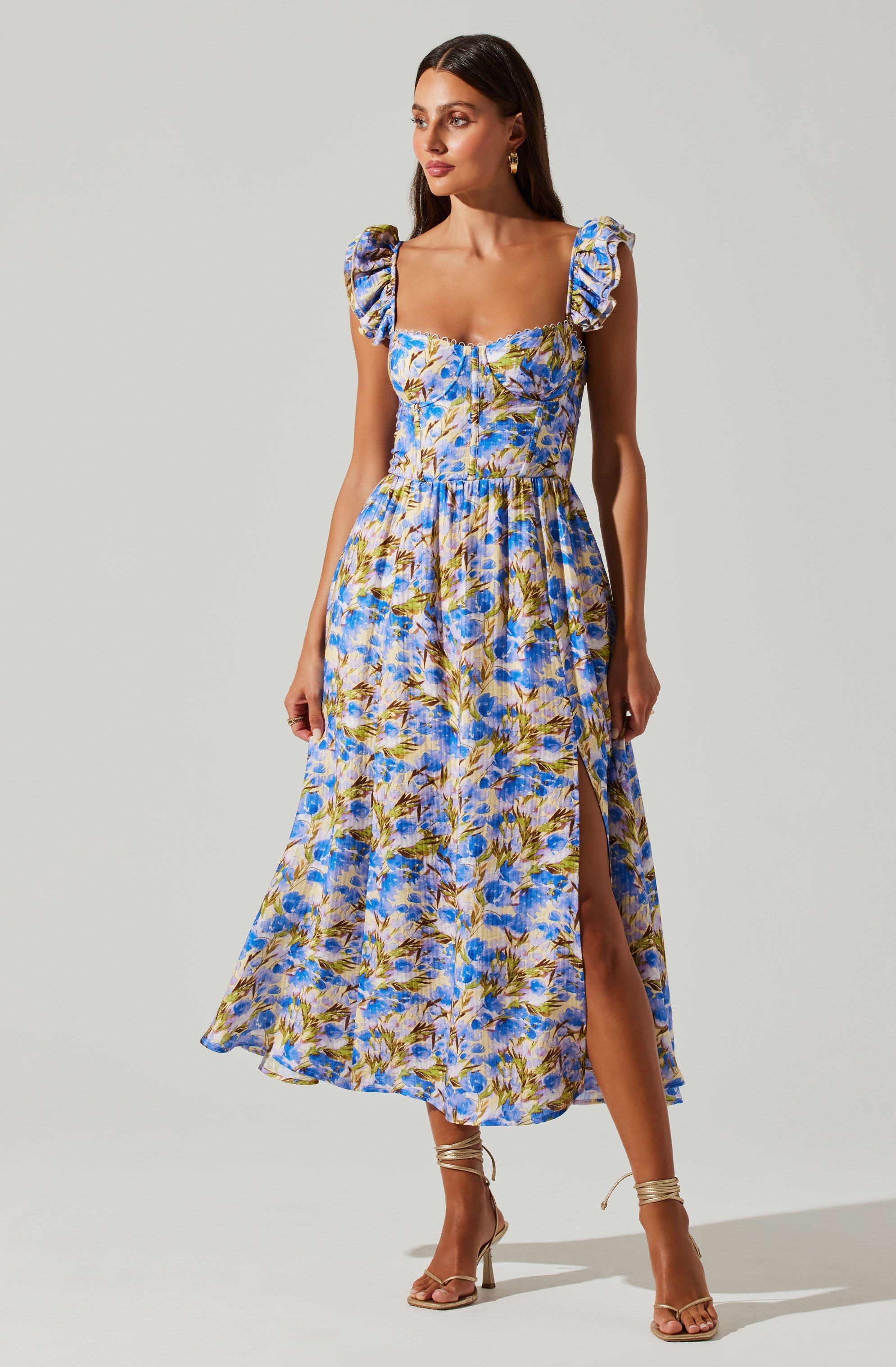 A woman is wearing a sleeveless, ankle-length dress with a floral pattern that includes shades of blue, purple, yellow, and green. The dress features ruffled shoulder straps, a fitted bodice, and a high slit on one side. She is also wearing strappy high-heeled sandals in a metallic gold color.