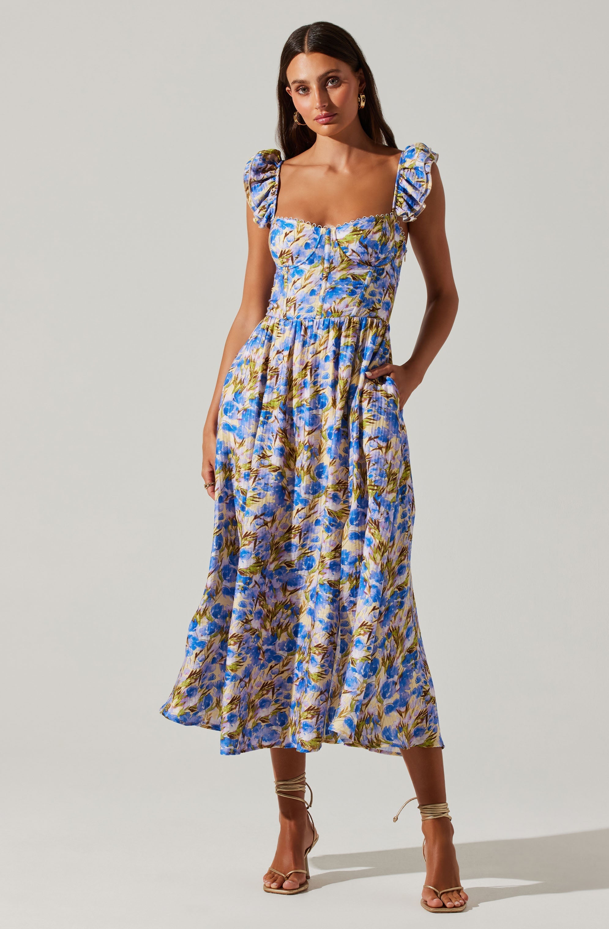A woman wearing a sleeveless, ankle-length dress with a floral pattern in shades of blue, purple, green, and yellow. The dress features ruffled shoulder straps and a fitted bodice with a slight sweetheart neckline. She pairs the dress with nude, open-toed high heels with ankle straps.