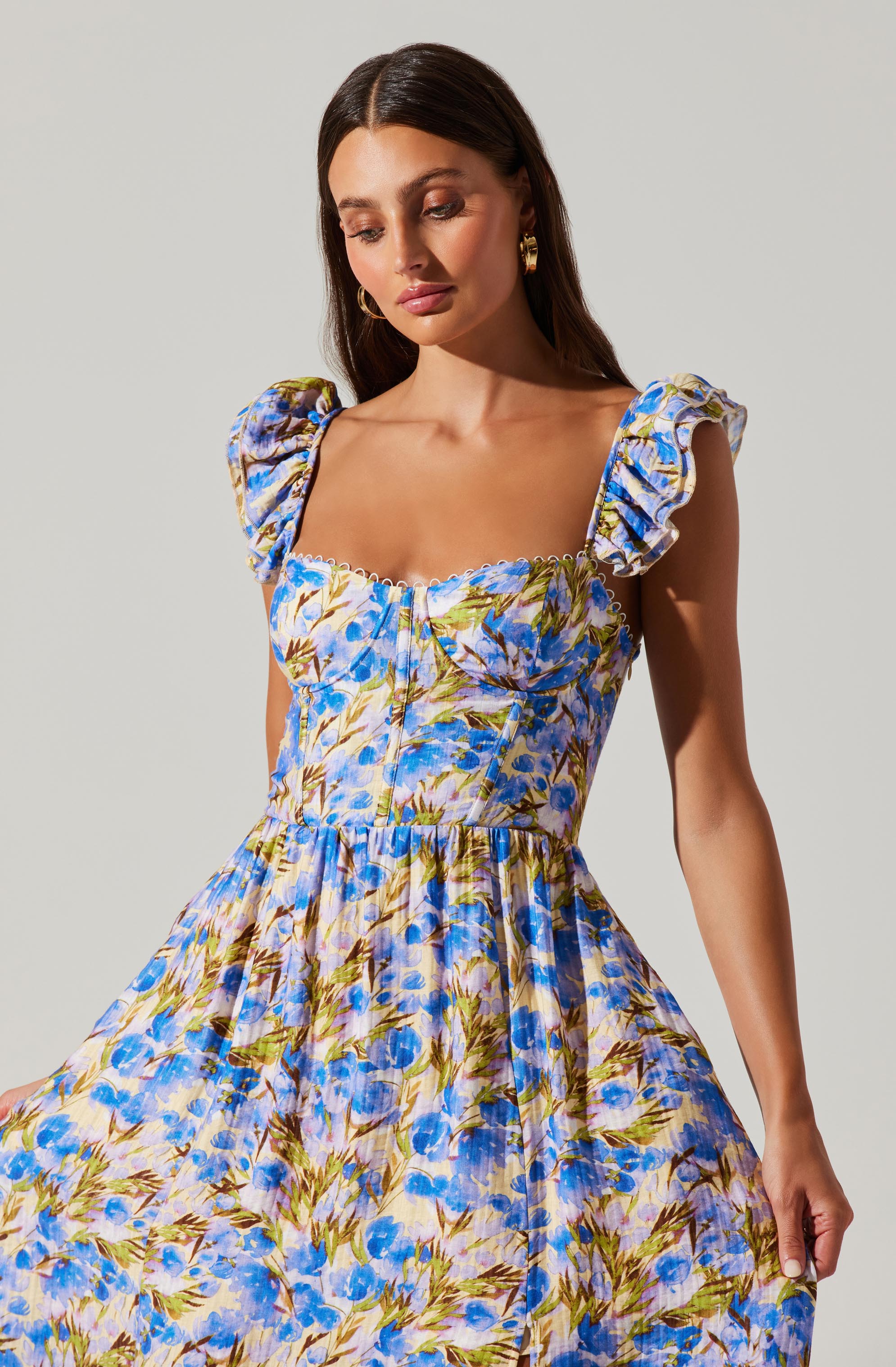 A woman is wearing a dress with a colorful floral pattern featuring shades of blue, yellow, and green. The dress has puffed, gathered short sleeves, a fitted bodice with a ruched design, and a gathered waistline that leads to a flowing skirt. The fabric appears lightweight and feminine.