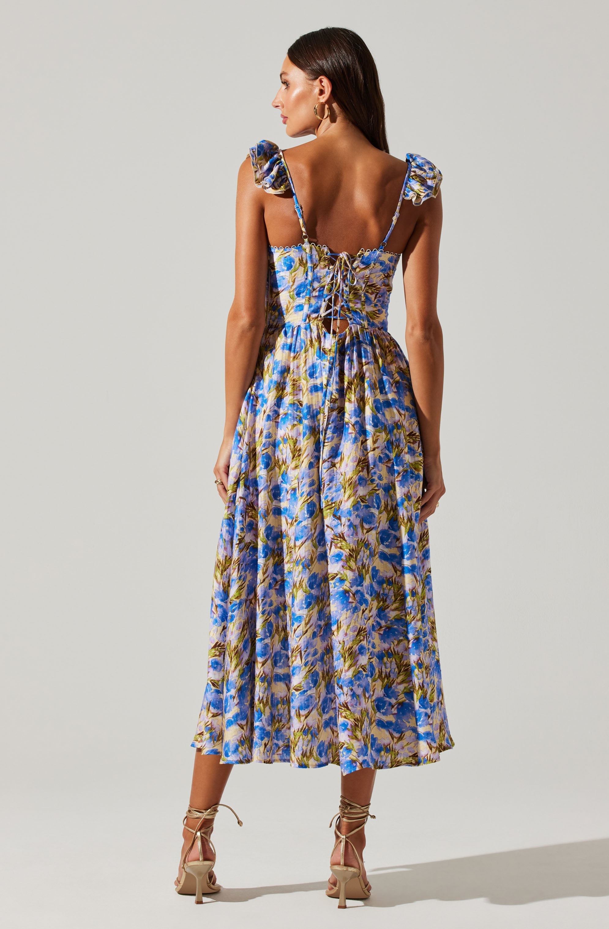A woman is wearing a long, flowy dress with a vibrant floral pattern in shades of blue, purple, and yellow. The dress features ruffled shoulder straps, a fitted waist with a lace-up back detail, and a full skirt that extends to her ankles. She pairs the dress with nude high-heeled sandals that have ankle ties.