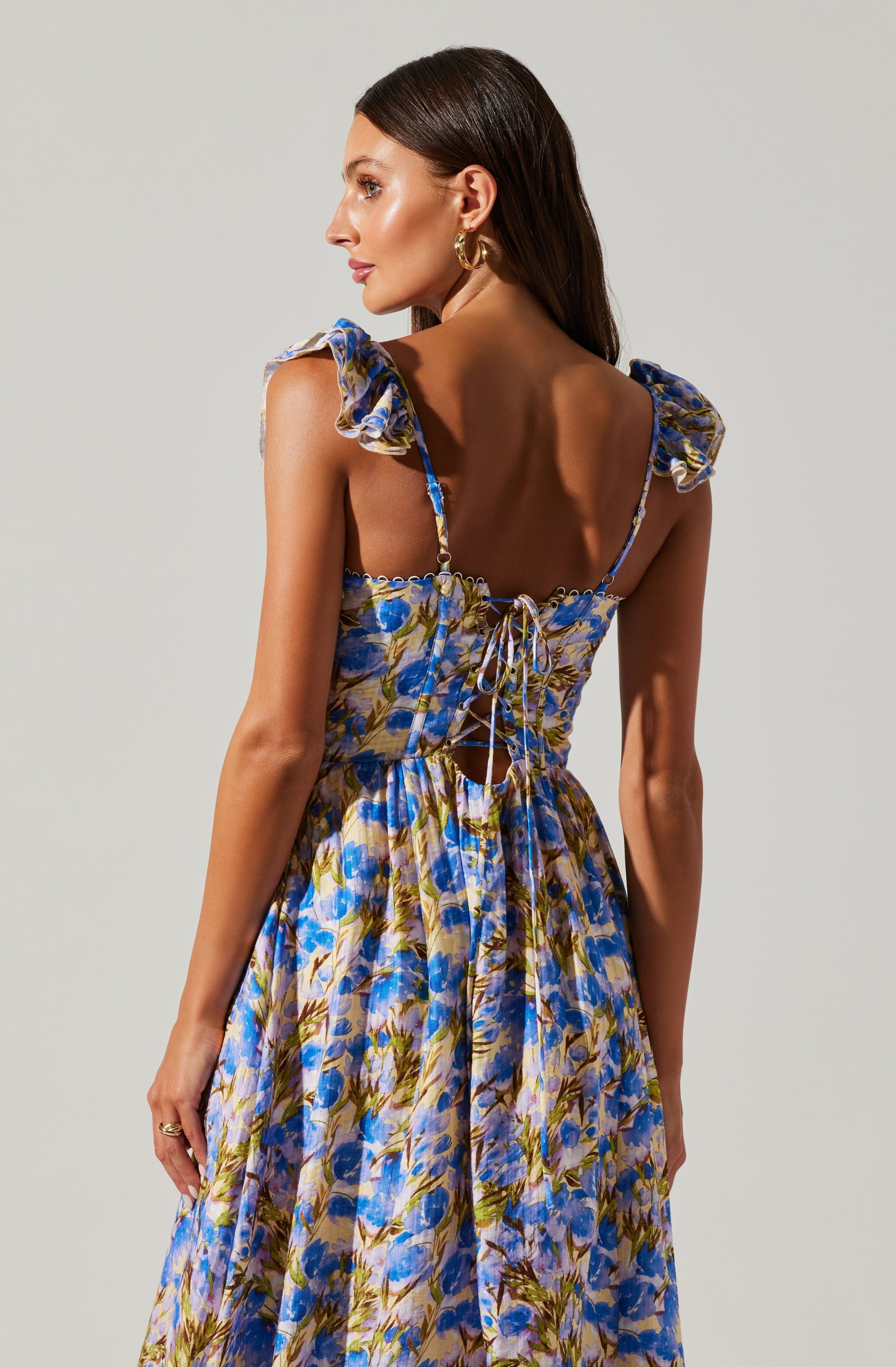 A woman wearing a sleeveless, floral dress with ruffled shoulder straps. The dress features a lace-up back with thin ties at the waist. The fabric has a colorful pattern of blue, purple, and green flowers.