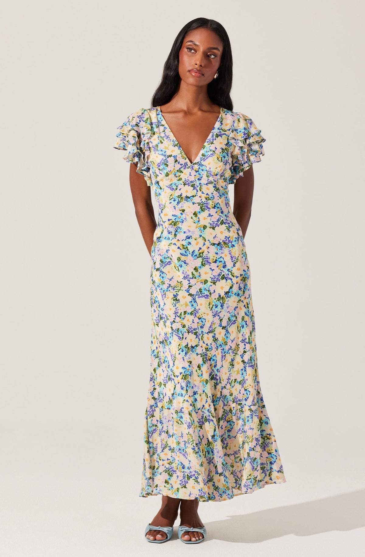 A woman is wearing a long, flowy dress with a floral pattern in shades of blue, purple, green, and beige on a light background. The dress features a V-neckline and short, ruffled sleeves.