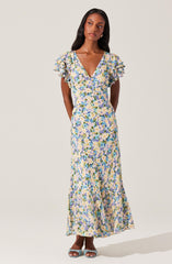 A woman is wearing a long, flowy dress with a floral pattern in shades of blue, purple, green, and beige on a light background. The dress features a V-neckline and short, ruffled sleeves.