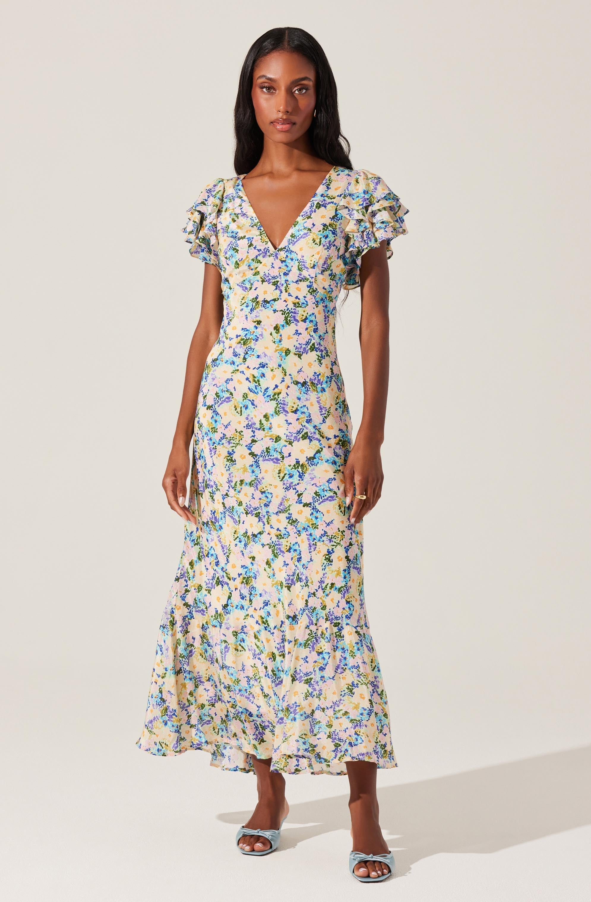 A woman is wearing a long, flowy dress with a floral pattern in shades of blue, yellow, and green on a light background. The dress features a V-neckline and short ruffled sleeves. She is also wearing a pair of open-toed heels with a bow detail.