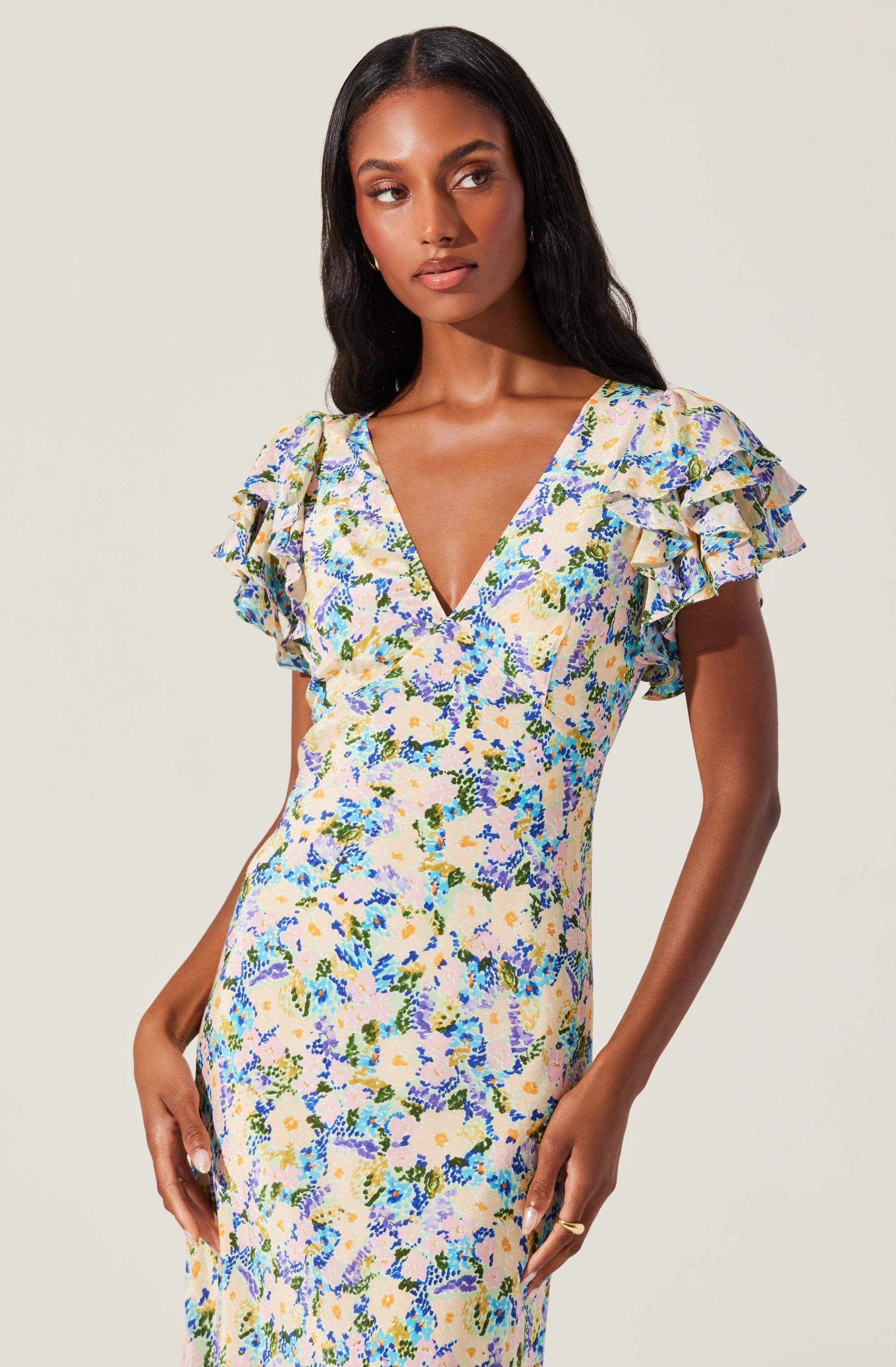 A woman is wearing a dress with a floral pattern that features shades of yellow, blue, green, and purple. The dress has a deep V neckline and short, ruffled sleeves.