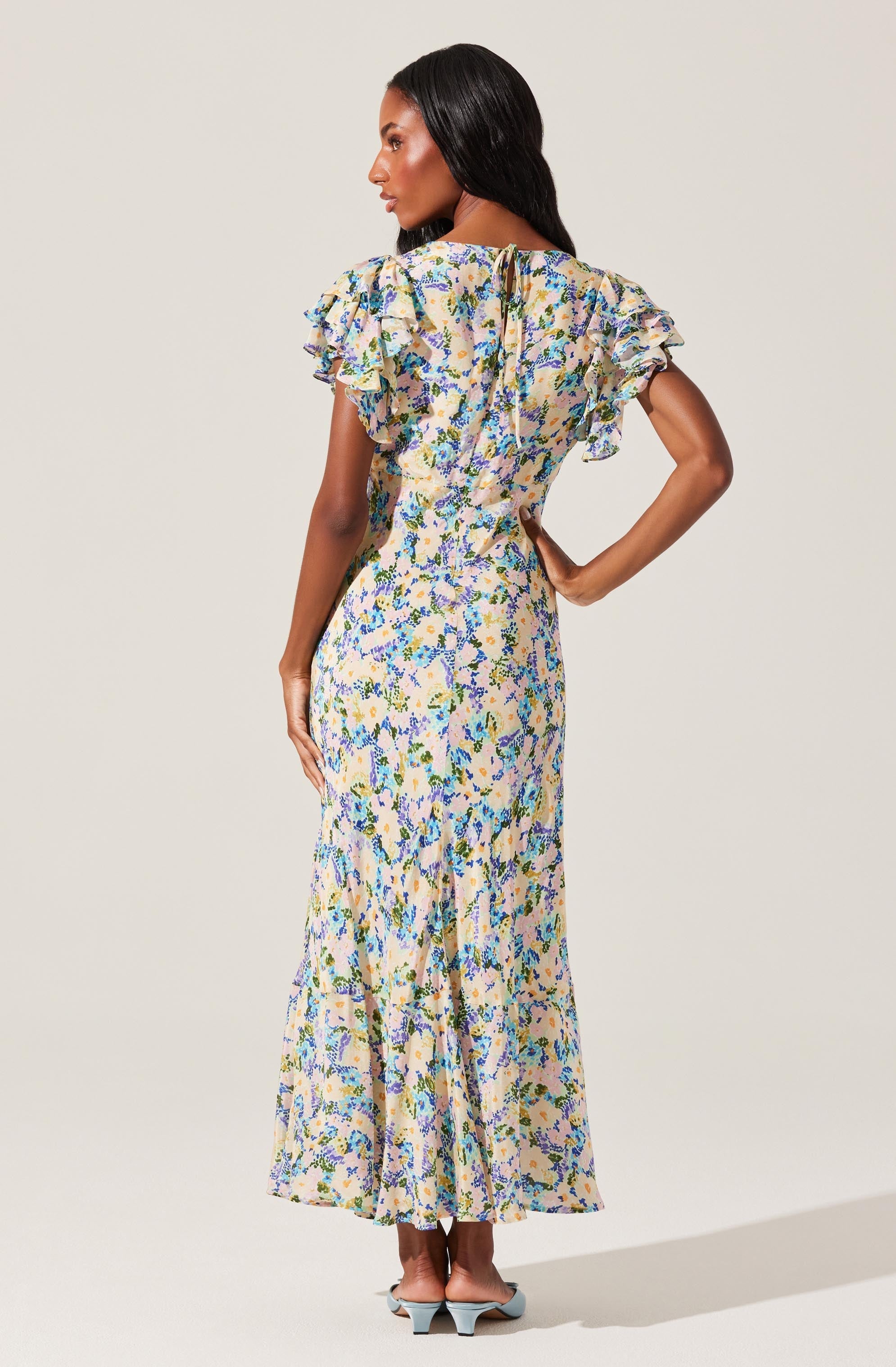 A woman is wearing a long, floral-patterned dress with ruffled short sleeves. The dress features a fitted waist and a flowing skirt that reaches the floor. The fabric has a light background with a multicolored floral design, primarily in shades of blue, green, yellow, and pink. She is also wearing light blue pointed-toe shoes with a low heel.