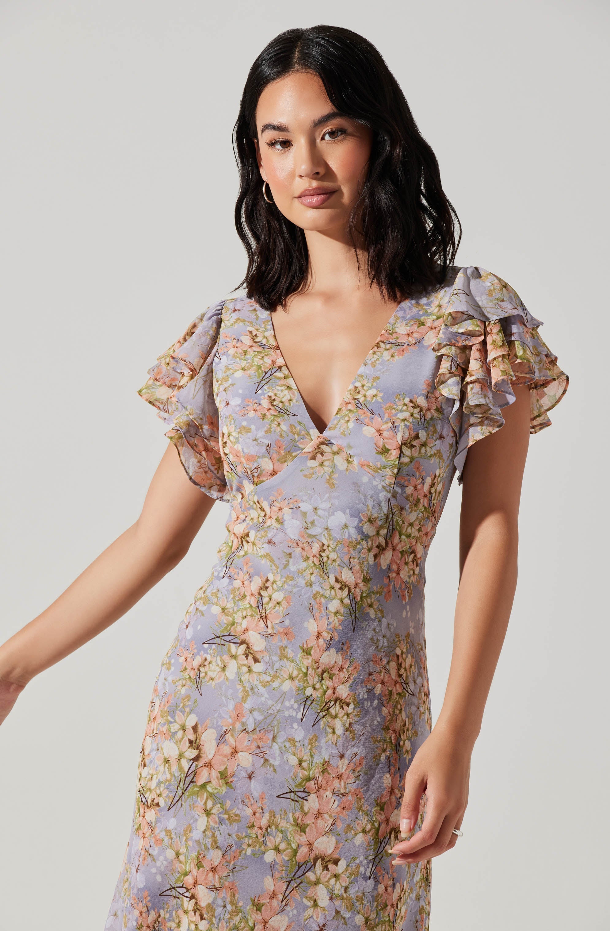 A woman is wearing a dress with a pastel purple background featuring a floral print in shades of pink, green, and cream. The dress has a V-neckline and short, ruffled sleeves made of a lightweight, semi-transparent fabric.