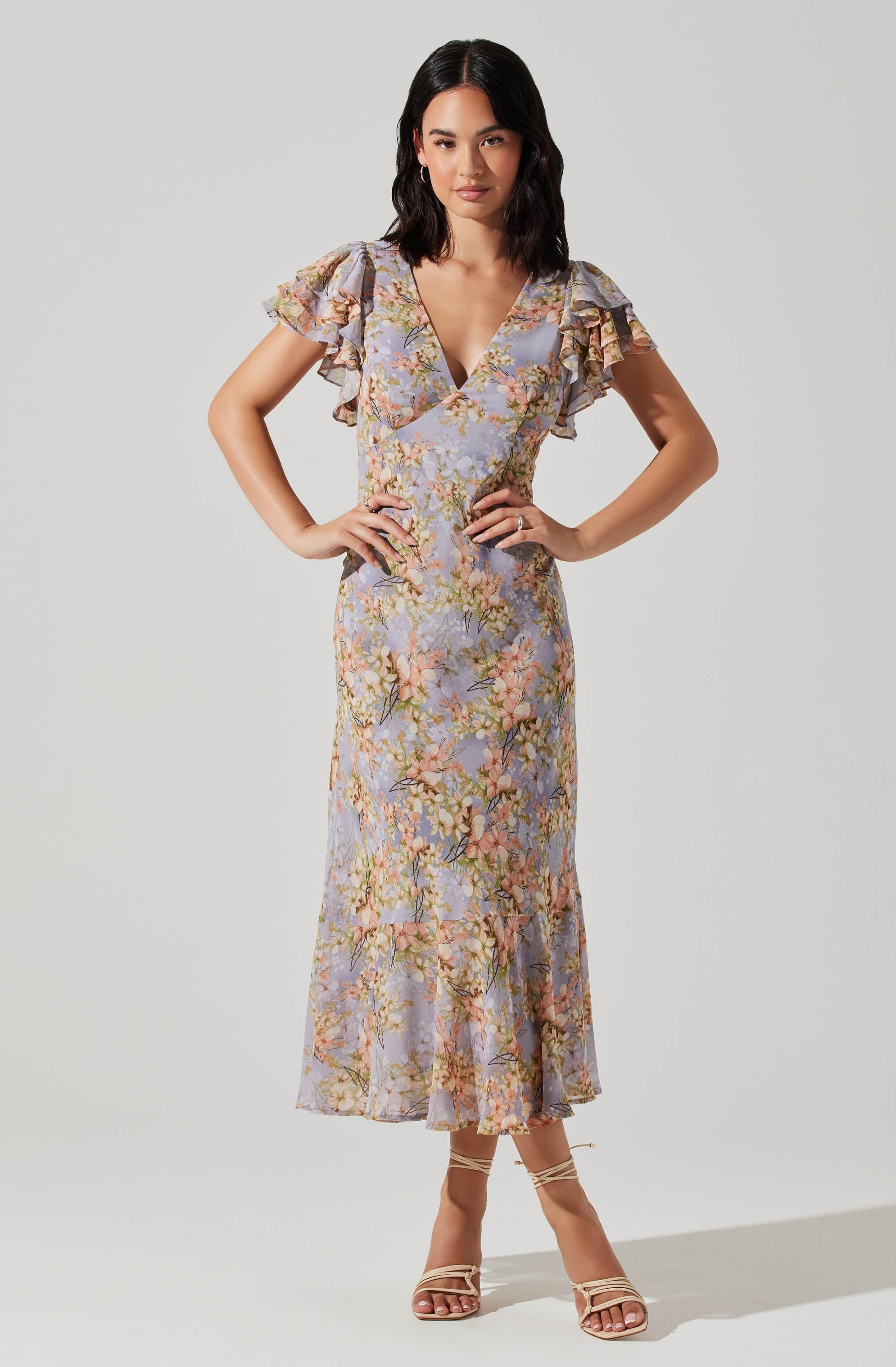 A woman is wearing a long, floral dress with a fitted waist and a ruffled V-neckline. The dress features short, ruffled sleeves and a tiered, ruffled hem that reaches mid-calf. The fabric has a pastel-colored floral pattern with shades of lavender, pink, green, and beige.