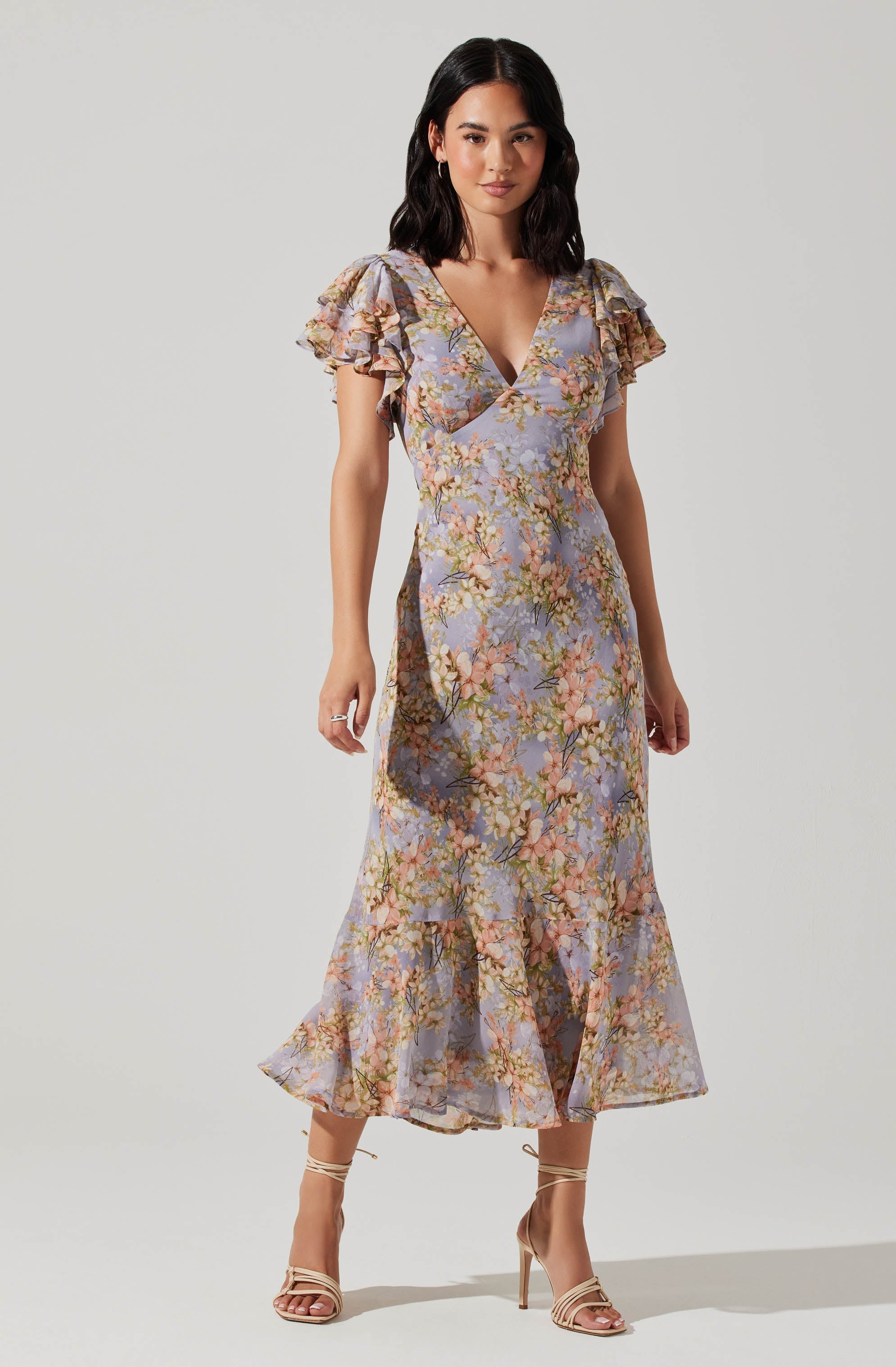 A woman is wearing a long, flowing dress with a floral pattern. The dress features short, ruffled sleeves, a V-neckline, and a ruffled hem that reaches above her ankles. The fabric has a soft, pastel background with multicolored flowers.