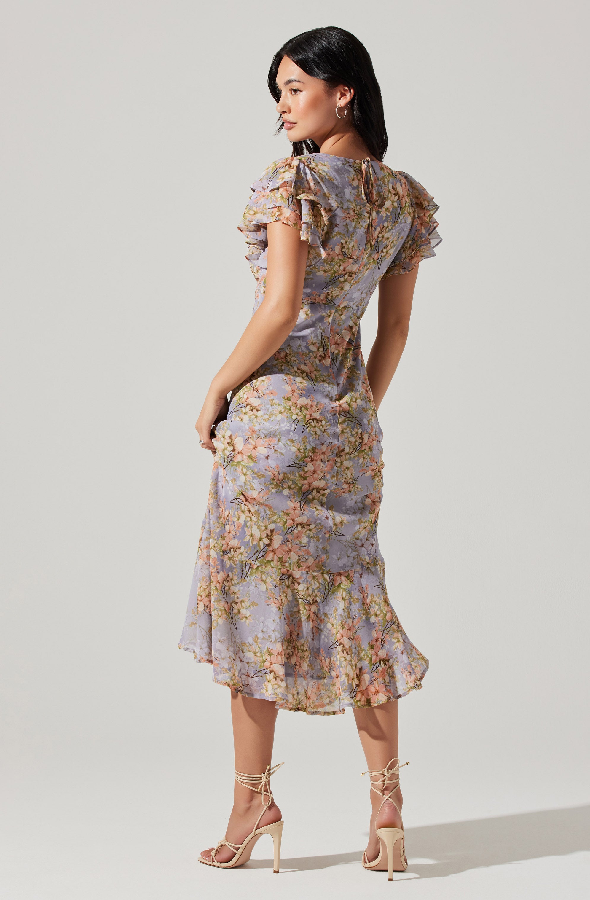 A woman is wearing a midi-length dress with a floral print in pastel shades, featuring ruffled short sleeves and a fitted waist. The dress has a sheer, lightweight fabric with a flowy silhouette. She pairs the dress with nude high-heeled sandals that have ankle ties.