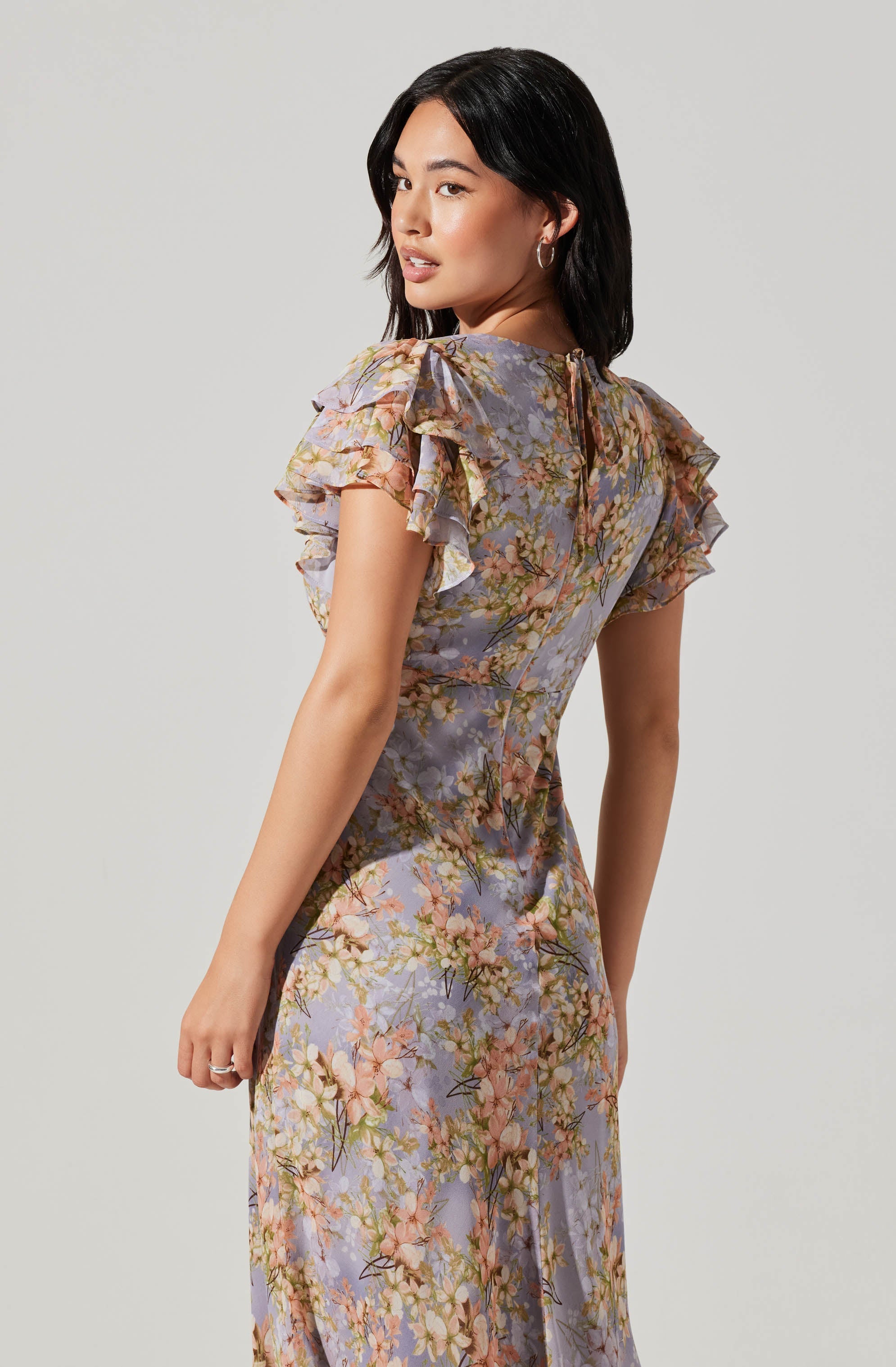 A woman is wearing a floral dress with a fitted silhouette. The dress features a pastel color palette with shades of lavender, pink, green, and beige. It has short, ruffled sleeves and a quarter-length zipper at the back. The fabric appears lightweight and slightly sheer, with a flowing skirt that reaches below the knees.