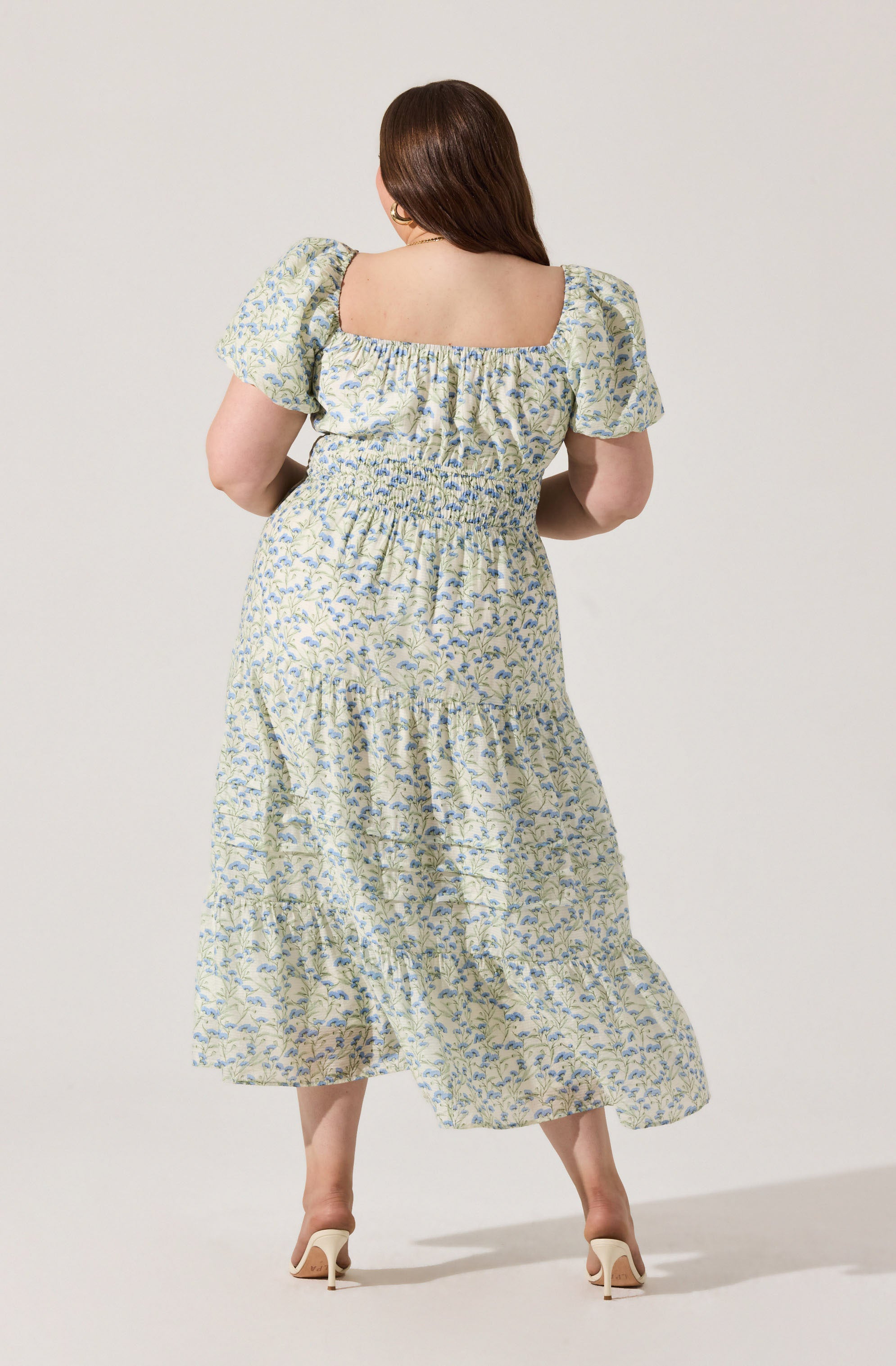 A woman is wearing a light-colored, floral-patterned dress with a square neckline and puffed short sleeves. The dress has a smocked waist and a tiered, flowing skirt that reaches mid-calf. She is also wearing white high-heeled shoes.