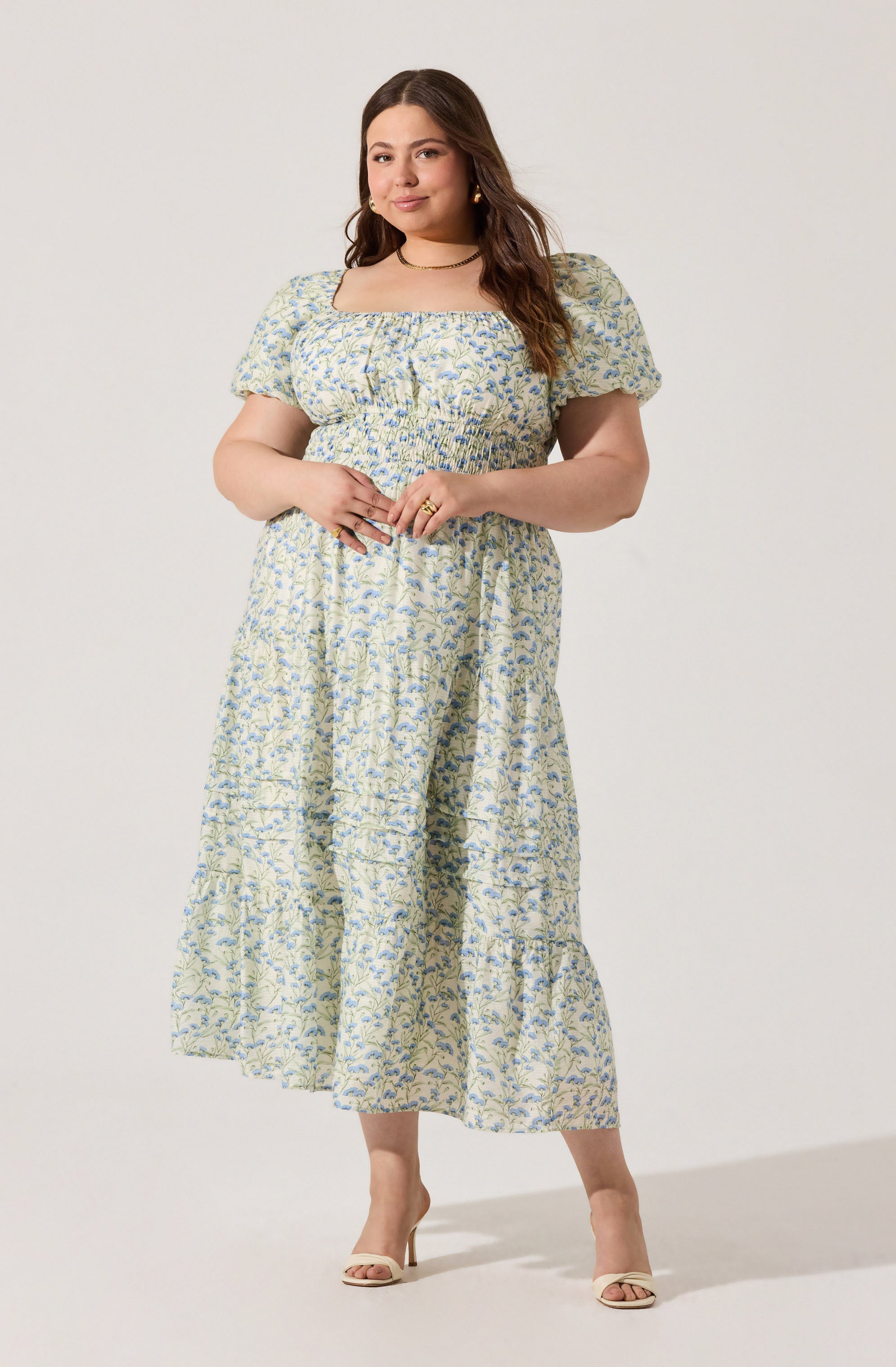 A woman is wearing a light-colored, floral-patterned dress with short puffed sleeves and a square neckline. The dress has a smocked waistband and a tiered, ankle-length skirt. She is also wearing beige open-toe heels, gold hoop earrings, and a delicate choker necklace.