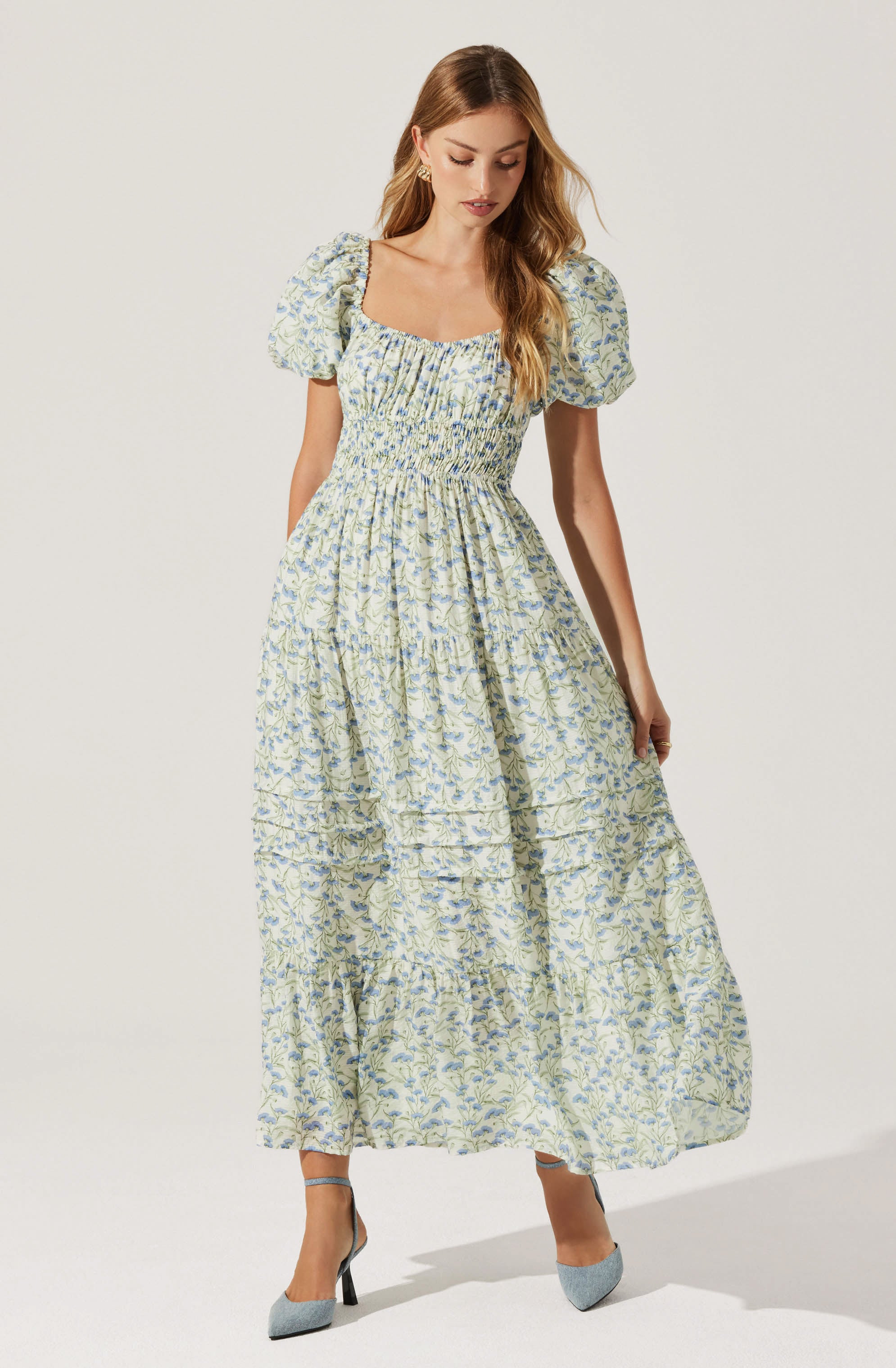 A woman is wearing a long, light-colored dress with a floral pattern. The dress features puffed short sleeves, a square neckline, and gathered fabric at the waist creating a slightly fitted bodice. The skirt is full and flowing, reaching down to her ankles. She is also wearing pointed-toe heels in a light gray color with black heels.
