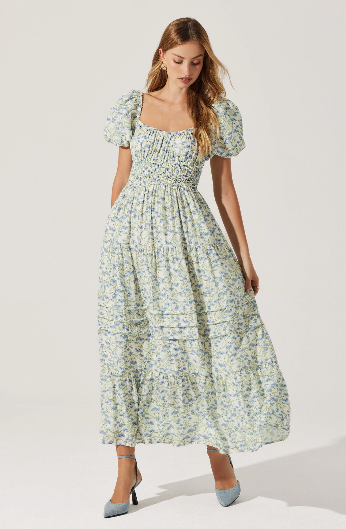 A woman is wearing a long, light-colored dress with a floral pattern. The dress features puffed short sleeves, a square neckline, and gathered fabric at the waist creating a slightly fitted bodice. The skirt is full and flowing, reaching down to her ankles. She is also wearing pointed-toe heels in a light gray color with black heels.
