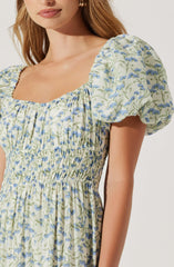 A woman is wearing a light-colored dress with a floral pattern featuring blue flowers and green leaves. The dress has a rounded neckline and puffed short sleeves. The fabric is gathered and smocked around the waist, creating a fitted bodice, and the skirt flows down from this gathered waist.