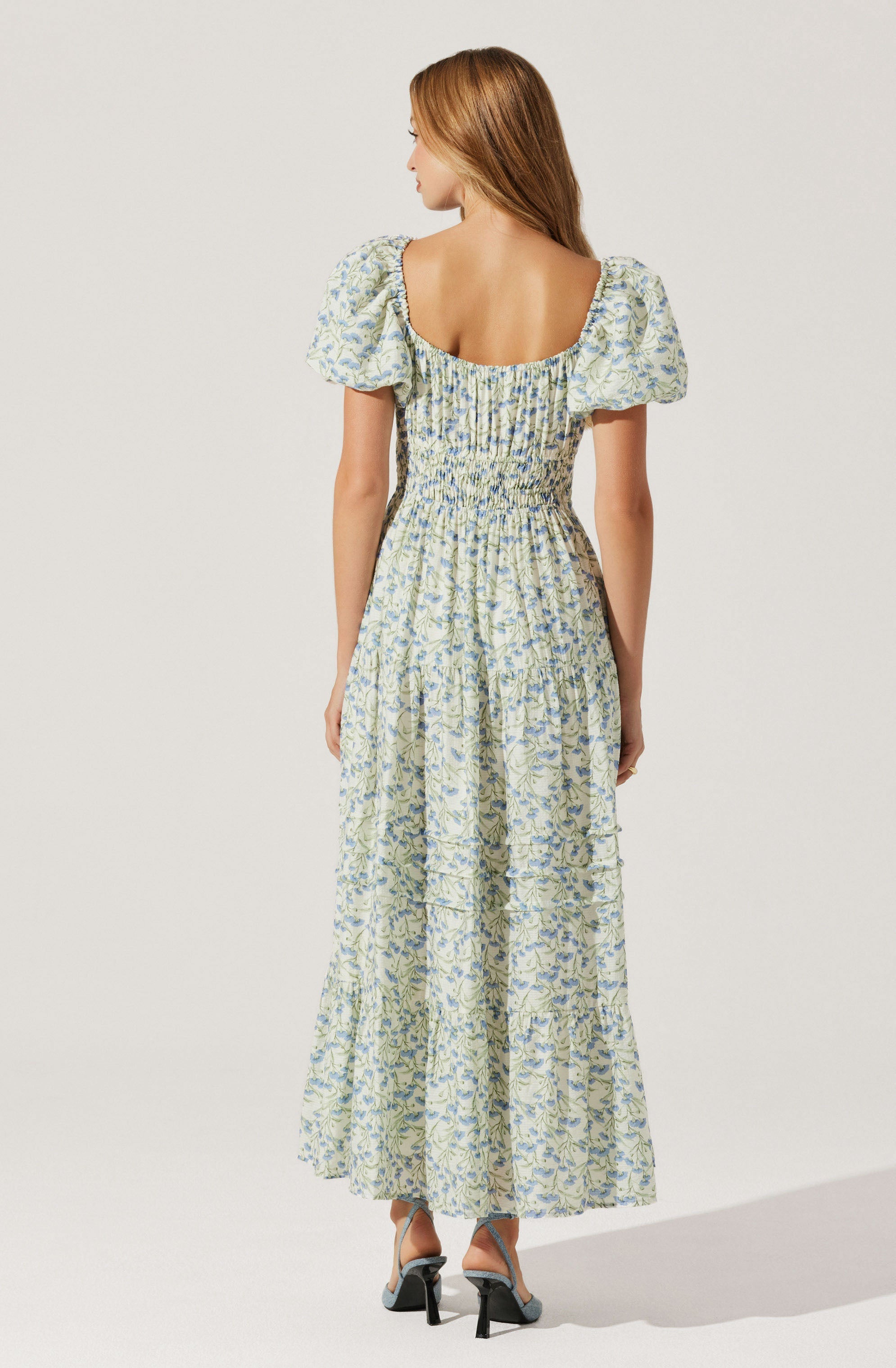 A woman is wearing a light-colored, floral-patterned maxi dress with puffed short sleeves and a wide, rounded neckline. The dress has a smocked waist and a flowing, tiered skirt that reaches the floor. She is also wearing high-heeled sandals.