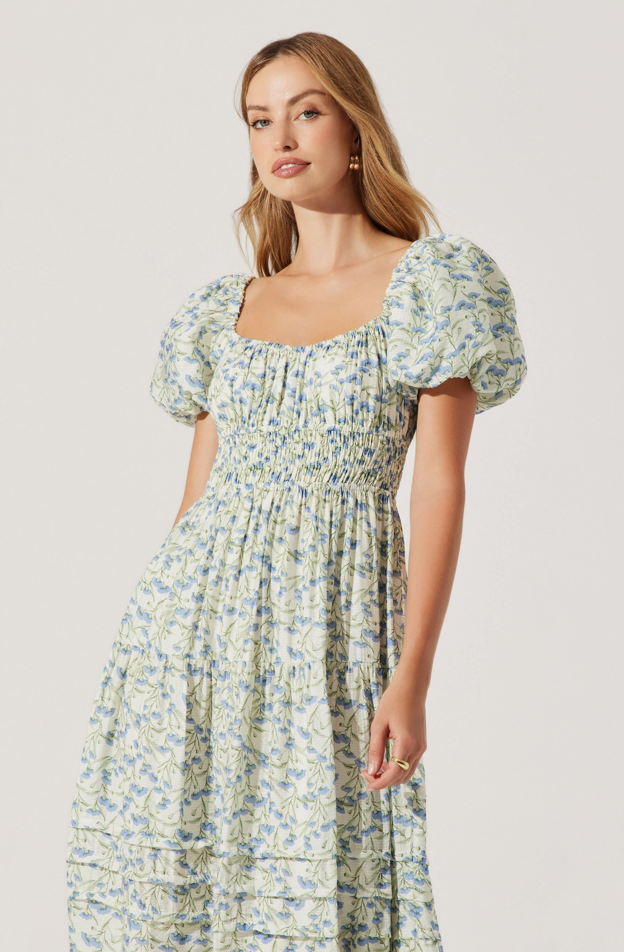 The woman is wearing a dress with a square neckline and short, puffed sleeves. The dress features a floral pattern in shades of blue and green on a light background. The bodice is fitted with shirring detail, and the skirt is long and flowing with a tiered design.