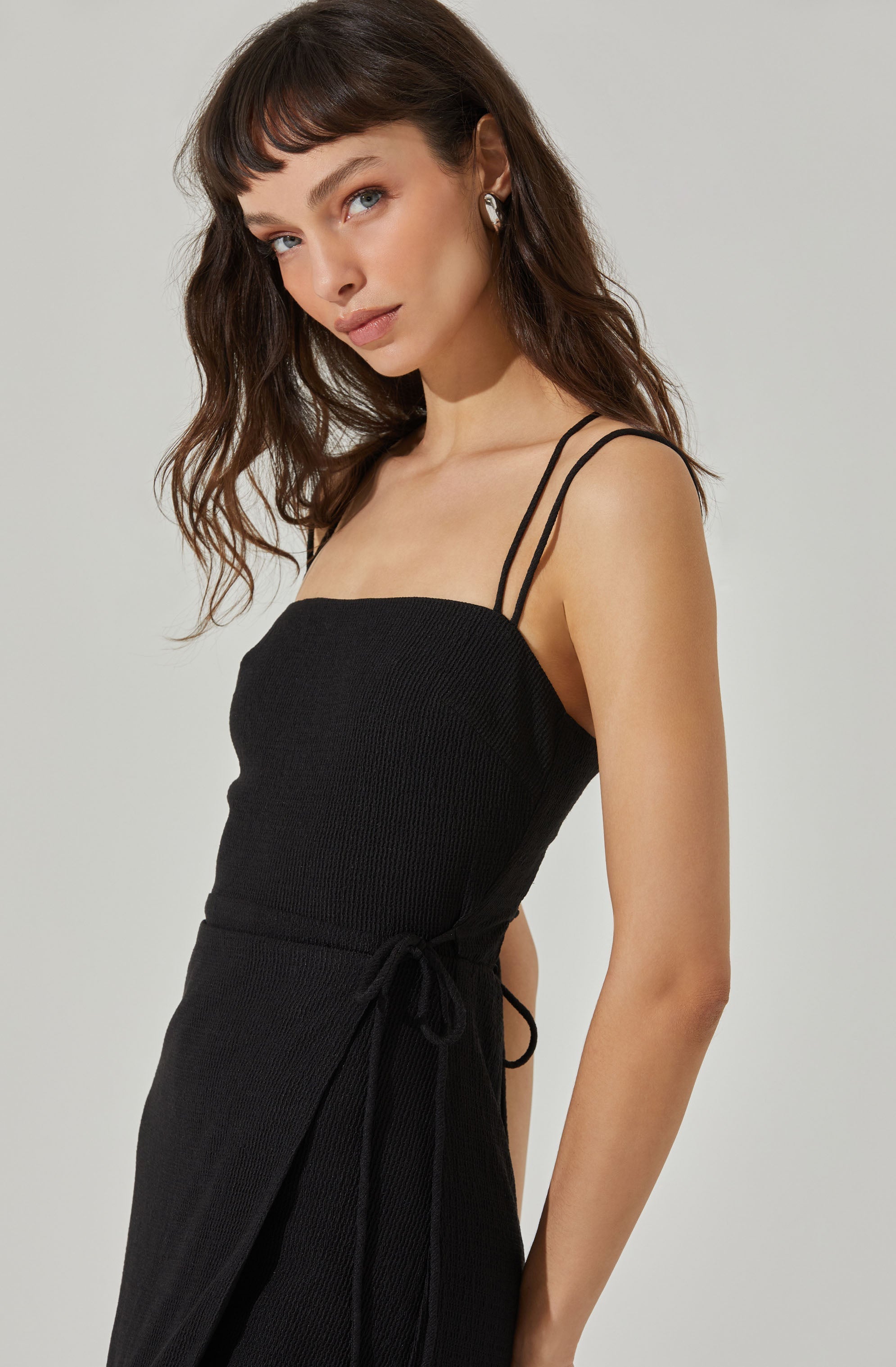 A young woman is wearing a black, sleeveless dress with thin double straps on each shoulder. The dress features a fitted bodice with a textured fabric and a waist tie on the side.