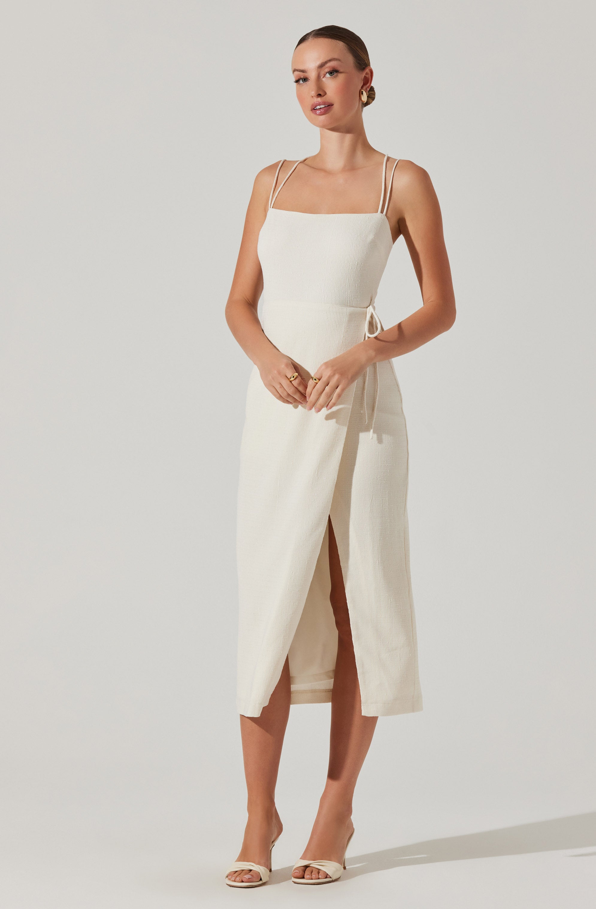 A woman is wearing a sleeveless, cream-colored dress with thin spaghetti straps. The dress has a fitted bodice and a long skirt with a front slit, allowing a glimpse of her legs. She is accessorizing with gold earrings and rings, and she is wearing neutral-colored high-heeled sandals.
