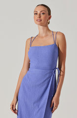 A woman is wearing a light blue, textured, sleeveless dress with thin spaghetti straps. The dress has a fitted bodice and a tie detail on the side.