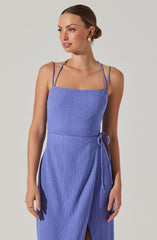A woman is wearing a sleeveless, light purple dress with thin double shoulder straps. The dress has a square neckline, a fitted waist, and a wrap style skirt with a front slit. The fabric appears textured and slightly ribbed.