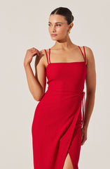 A woman is wearing a fitted, sleeveless red dress with thin double straps on each shoulder. The dress features a slit on one side and a drawstring detail at the waist.