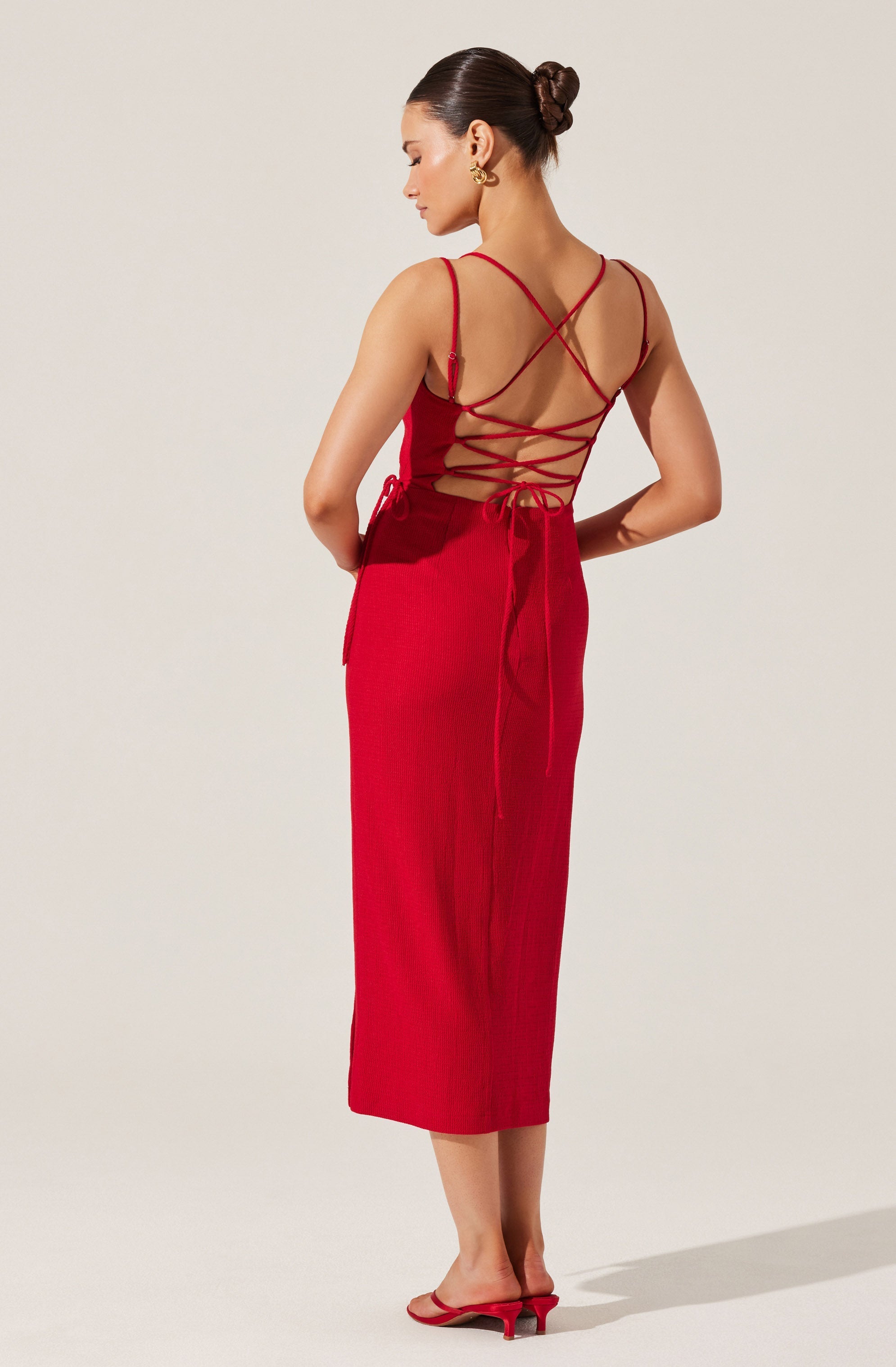 A woman is wearing a long, sleeveless red dress with a fitted bodice and a straight skirt that reaches mid-calf. The back of the dress features thin spaghetti straps and crisscrossing decorative ties that lace up along the open back, which is partially exposed. She has her hair styled in a sleek bun and is accessorized with large, gold hoop earrings. She is wearing matching red, open-toed heeled sandals.