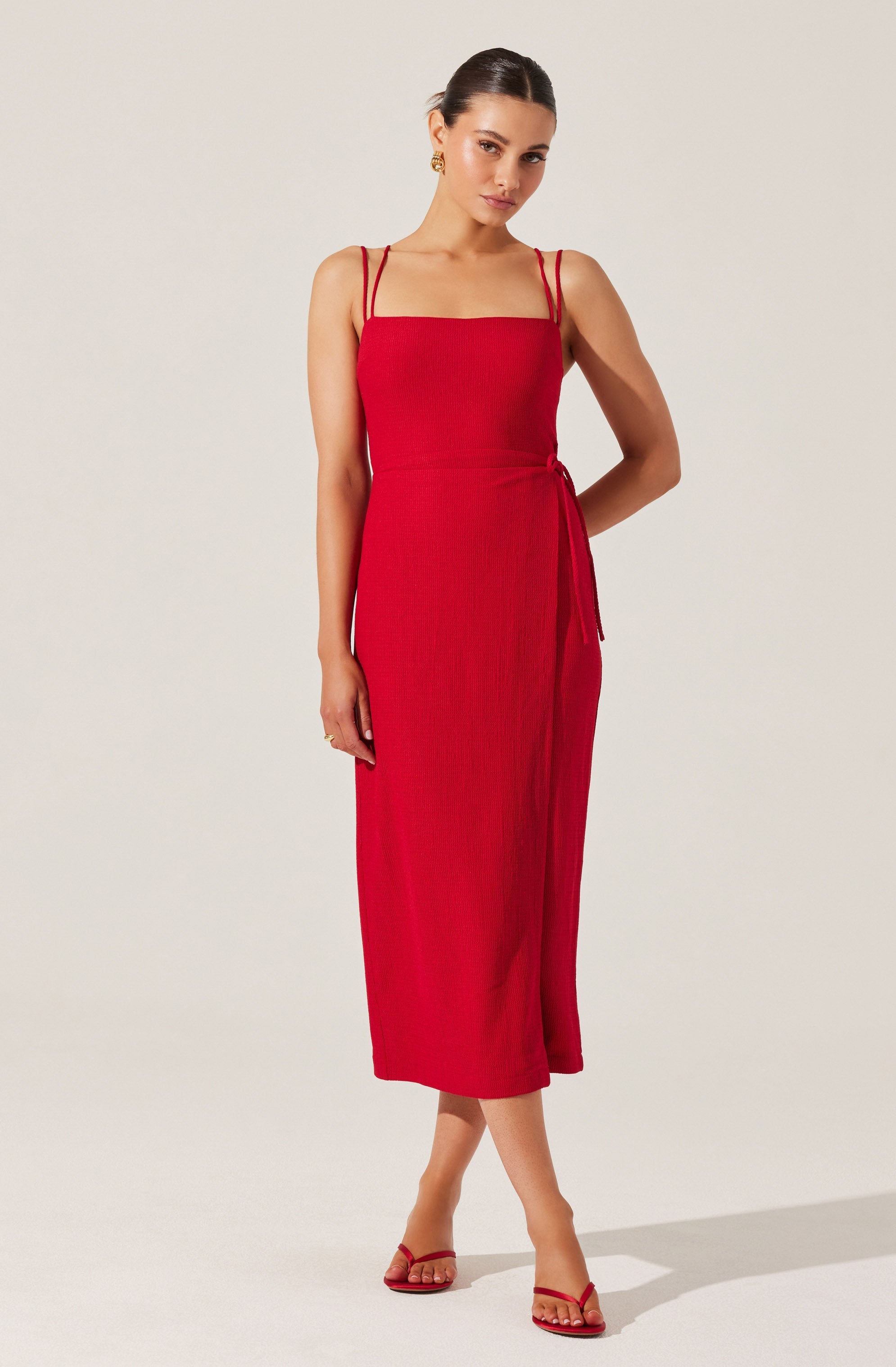 A woman is wearing a sleeveless, red, midi-length dress with thin double straps and a side tie detail. She is accessorized with gold earrings and red strappy high heels.