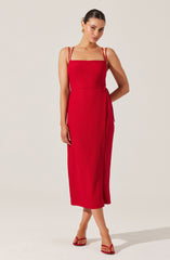 A woman is wearing a sleeveless, red, midi-length dress with thin double straps and a side tie detail. She is accessorized with gold earrings and red strappy high heels.