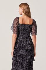 A woman is wearing a black dress with a floral pattern featuring small purple and green flowers. The dress has a square neckline and short, sheer puff sleeves. The fabric is lightweight and slightly sheer, with a smocked bodice and tiered ruffle skirt.