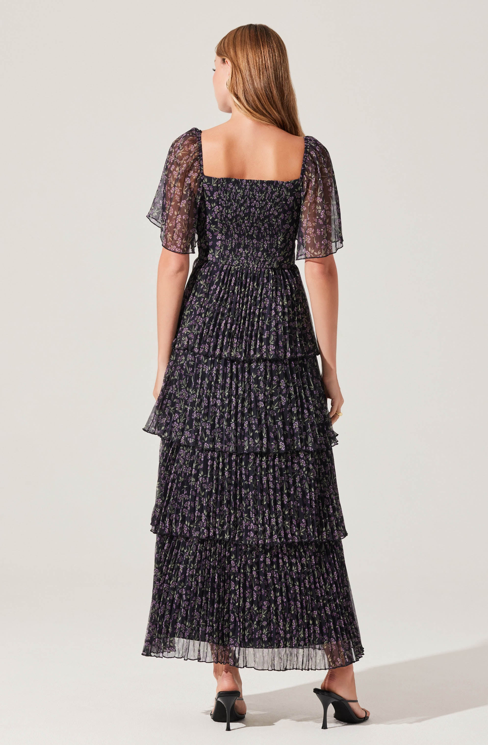 A woman is wearing a black floor-length dress with a small purple floral print. The dress features short, sheer puff sleeves made of a lightweight, semi-transparent fabric. The bodice is fitted with a square neckline. The skirt has multiple ruffled tiers, adding a layered, textured look. She is also wearing black high-heeled sandals.