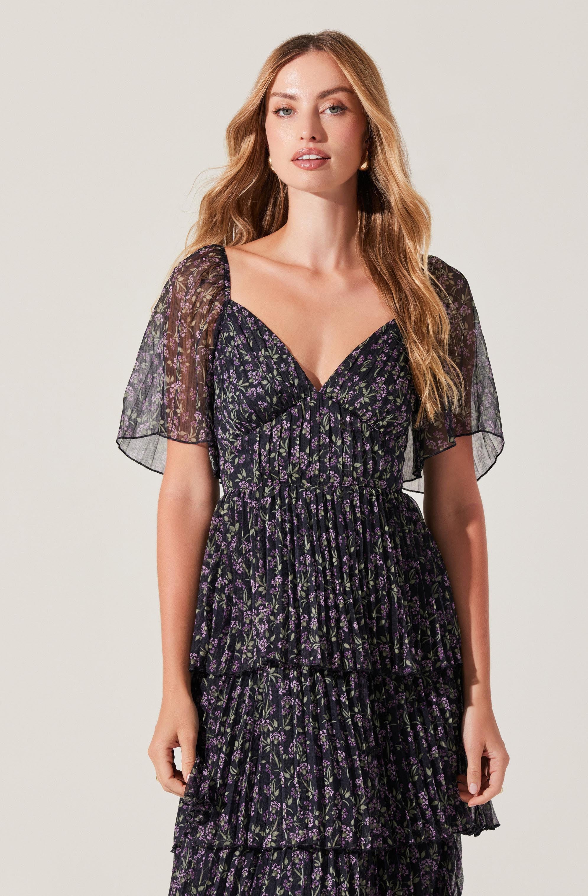 A woman is wearing a black dress with a small purple floral pattern. The dress has a deep V neckline, short sheer puff sleeves, and a fitted bodice with gathered fabric. The skirt of the dress features multiple tiers with ruffled edges.