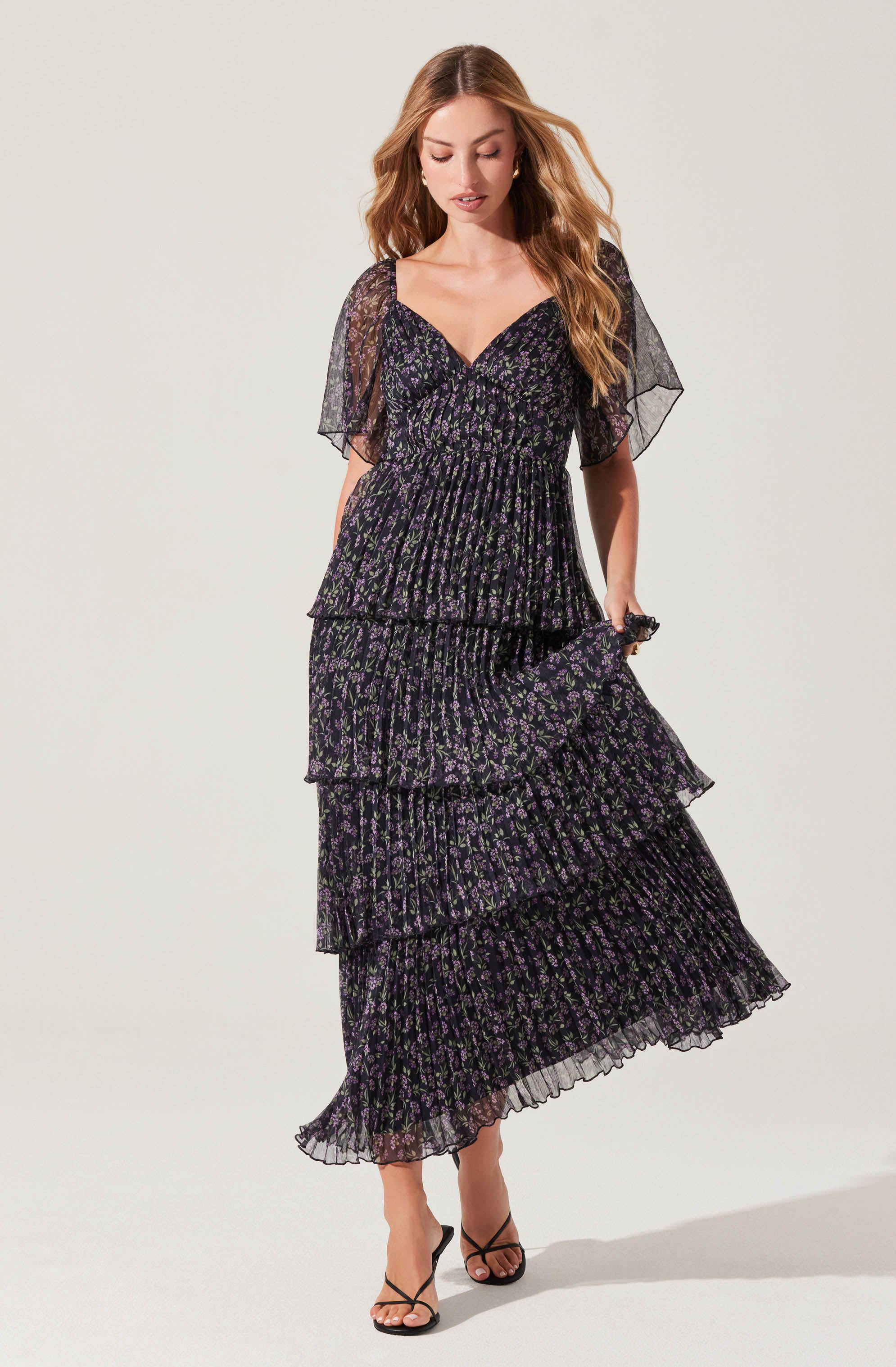 A woman is wearing a black maxi dress with purple and green floral patterns. The dress features a V-neckline, short sheer sleeves, and a tiered, ruffled skirt.