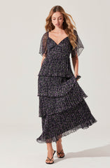 A woman is wearing a black maxi dress with purple and green floral patterns. The dress features a V-neckline, short sheer sleeves, and a tiered, ruffled skirt.