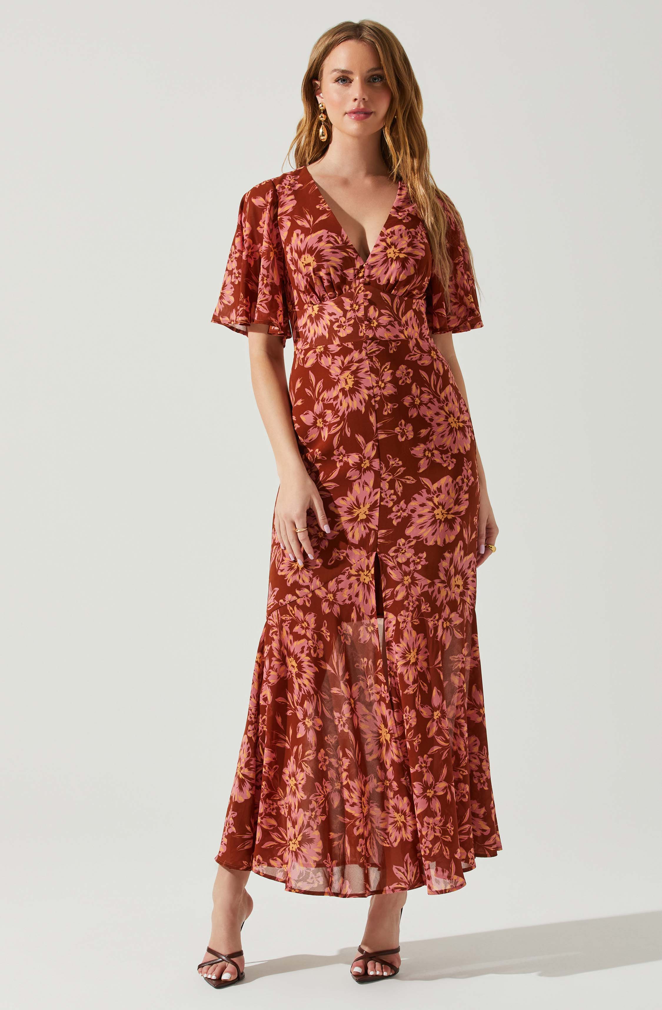 A woman is wearing a long, flowing, floral dress with shades of pink and orange on a dark reddish-brown background. The dress has short, ruffled sleeves and a V-neckline. The fabric features a large floral pattern, and the dress appears semi-sheer with a slit down the front. She is also wearing black strappy high-heeled sandals and statement earrings.