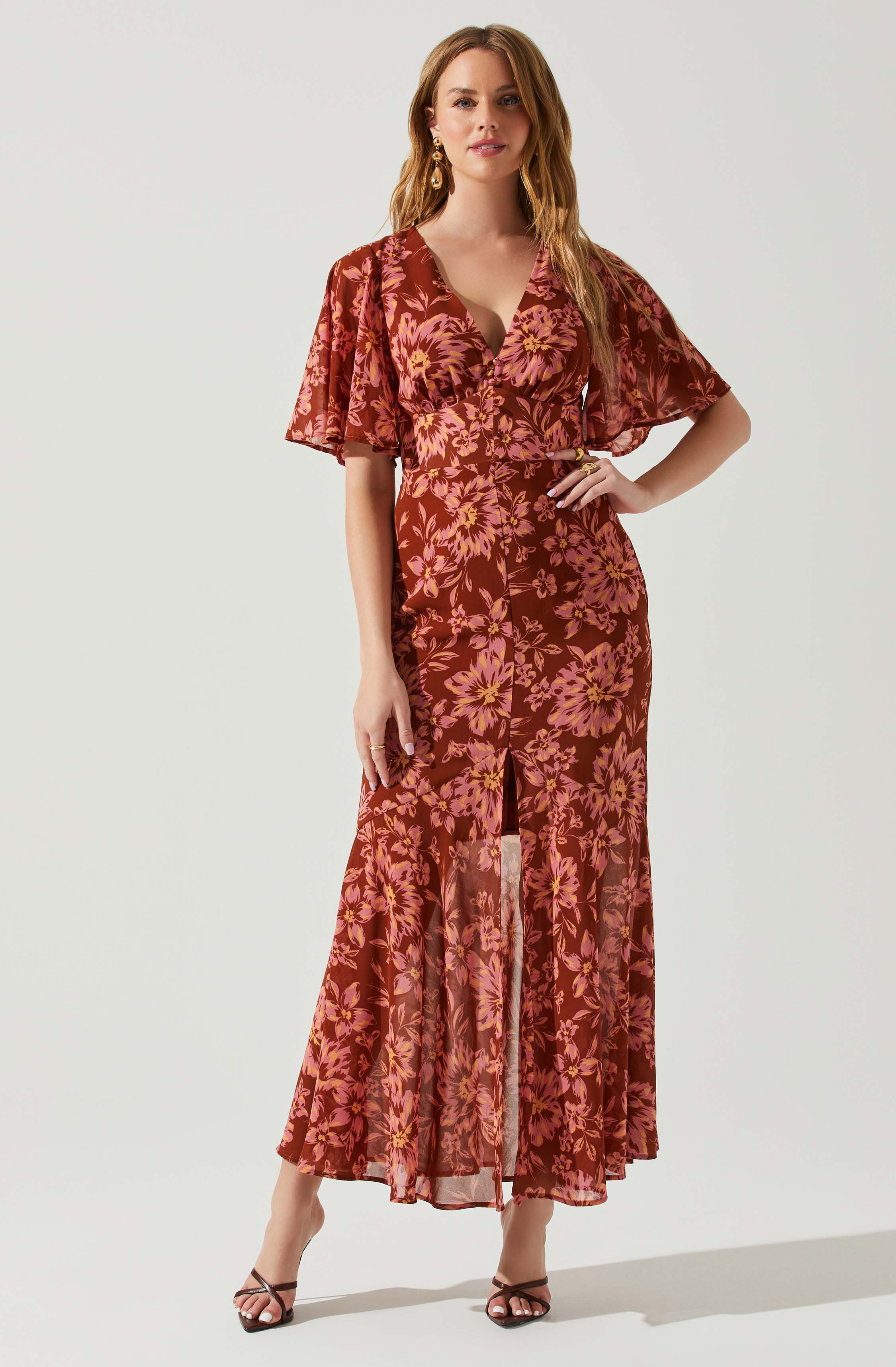 A woman wearing a long, flowing dress with a floral pattern in shades of pink and rust. The dress features a fitted waist, a deep V neckline, short flutter sleeves, and a front slit. She is accessorized with gold earrings and rings, and is standing in a relaxed pose with one hand on her hip.