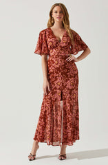 A woman wearing a long, flowing dress with a floral pattern in shades of pink and rust. The dress features a fitted waist, a deep V neckline, short flutter sleeves, and a front slit. She is accessorized with gold earrings and rings, and is standing in a relaxed pose with one hand on her hip.