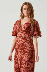 A woman wearing a flowing, floral-patterned dress with short, puffed sleeves. The dress has a deep V neckline, and the pattern features large pink and yellow flowers on a dark red background. She is accessorized with gold drop earrings and a gold ring on her right hand.