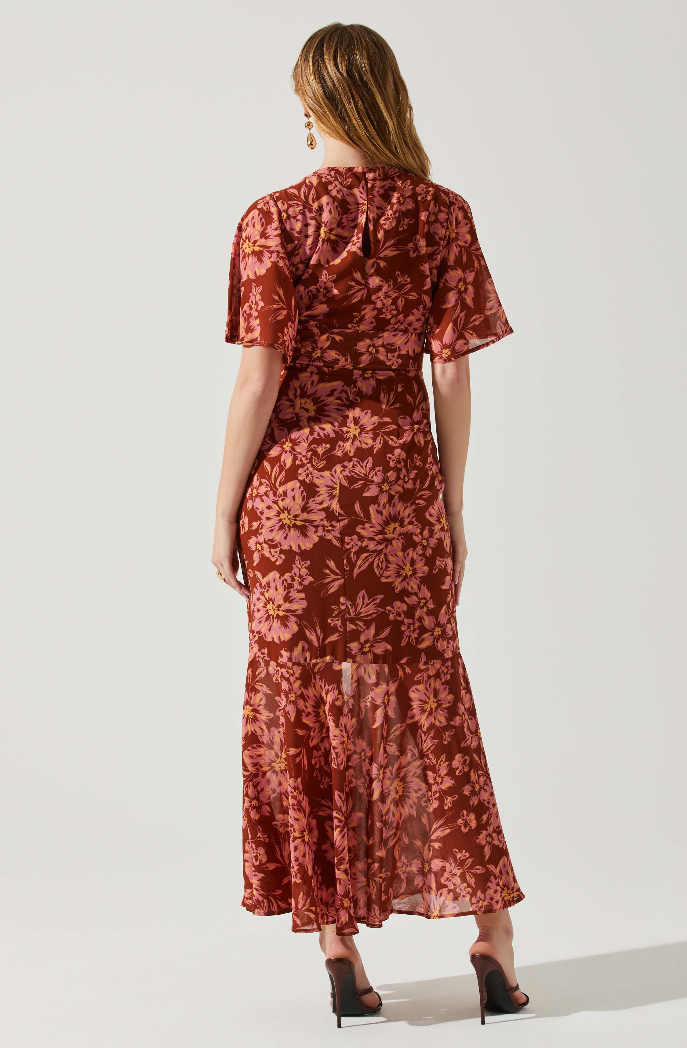A woman is wearing a long, flowing dress with a floral pattern in shades of pink and purple on a dark red background. The dress features short, puffed sleeves and a fitted waist, with a sheer, ruffled hem at the bottom. She has on high-heeled, open-toed shoes and is accessorized with dangling earrings.
