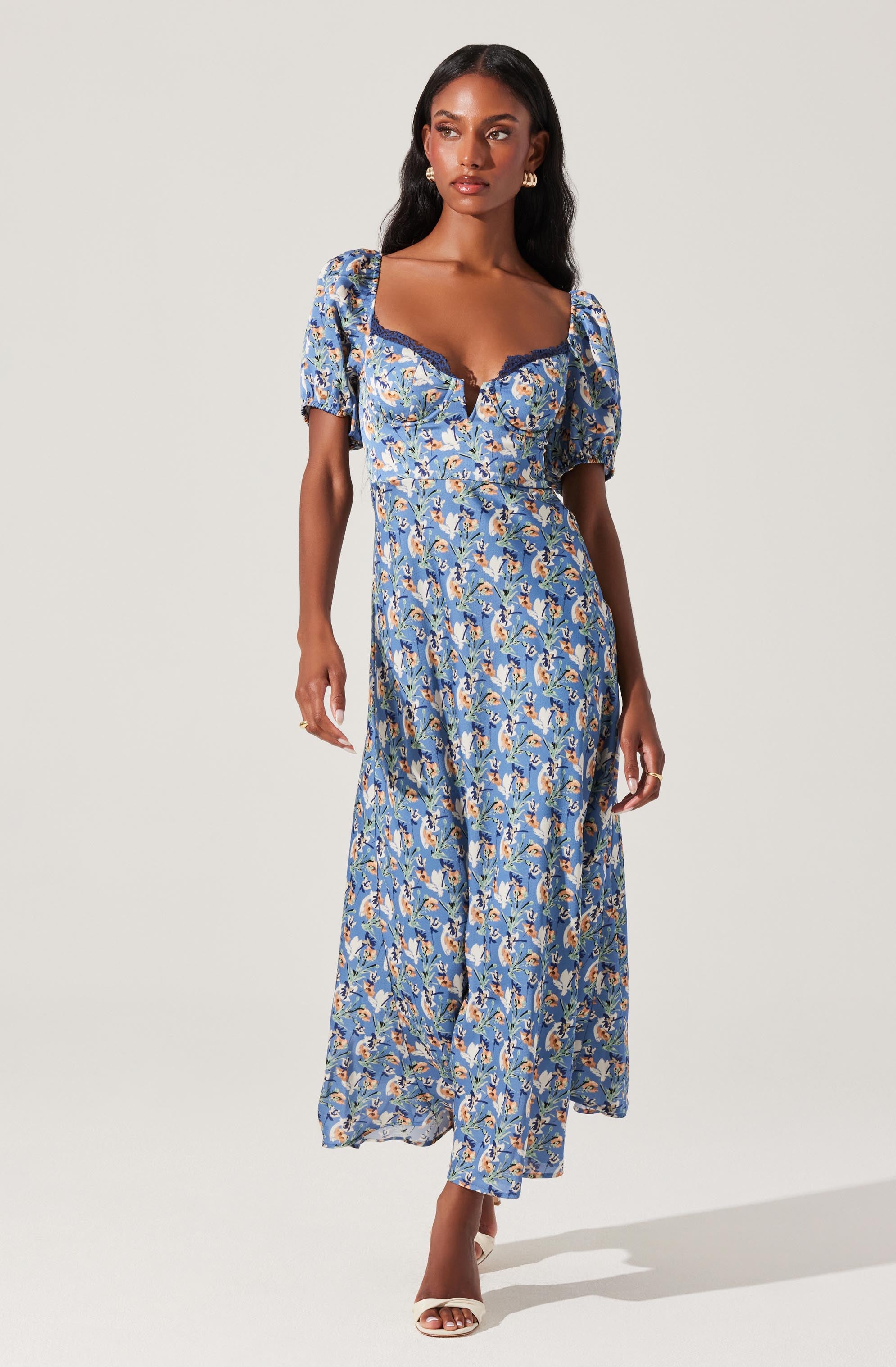 A woman is wearing a long, flowy dress with a blue floral pattern. The dress features puffed short sleeves, a sweetheart neckline with dark lace trim, and a fitted waist that flows into a full-length skirt. She is also wearing open-toe white heels and has long, dark hair styled in soft waves.