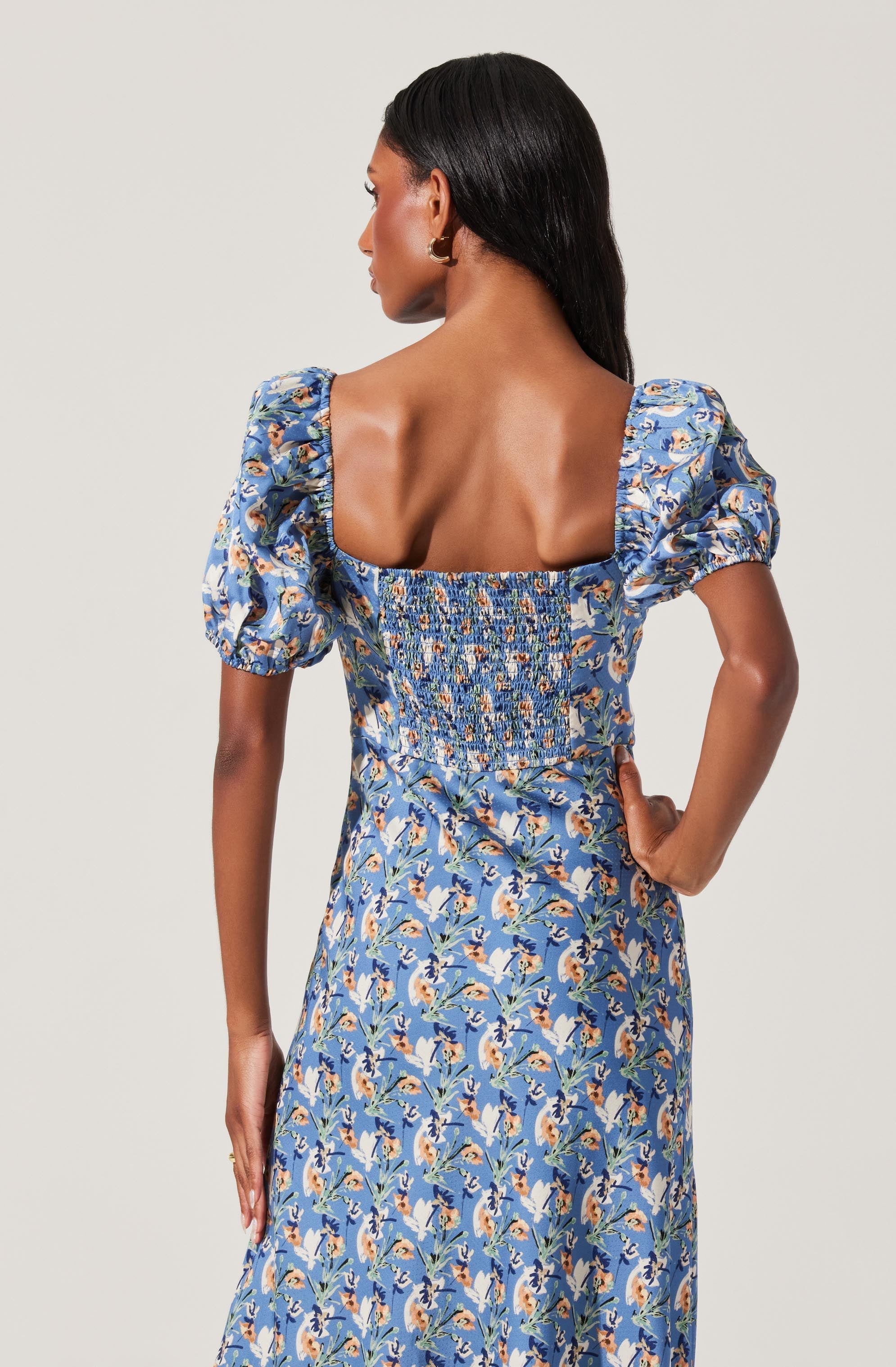 A woman is wearing a blue floral dress with short puffed sleeves and a fitted, smocked bodice. The dress features a pattern of white and peach flowers with green leaves and has a square neckline. The fabric appears lightweight with a soft drape.
